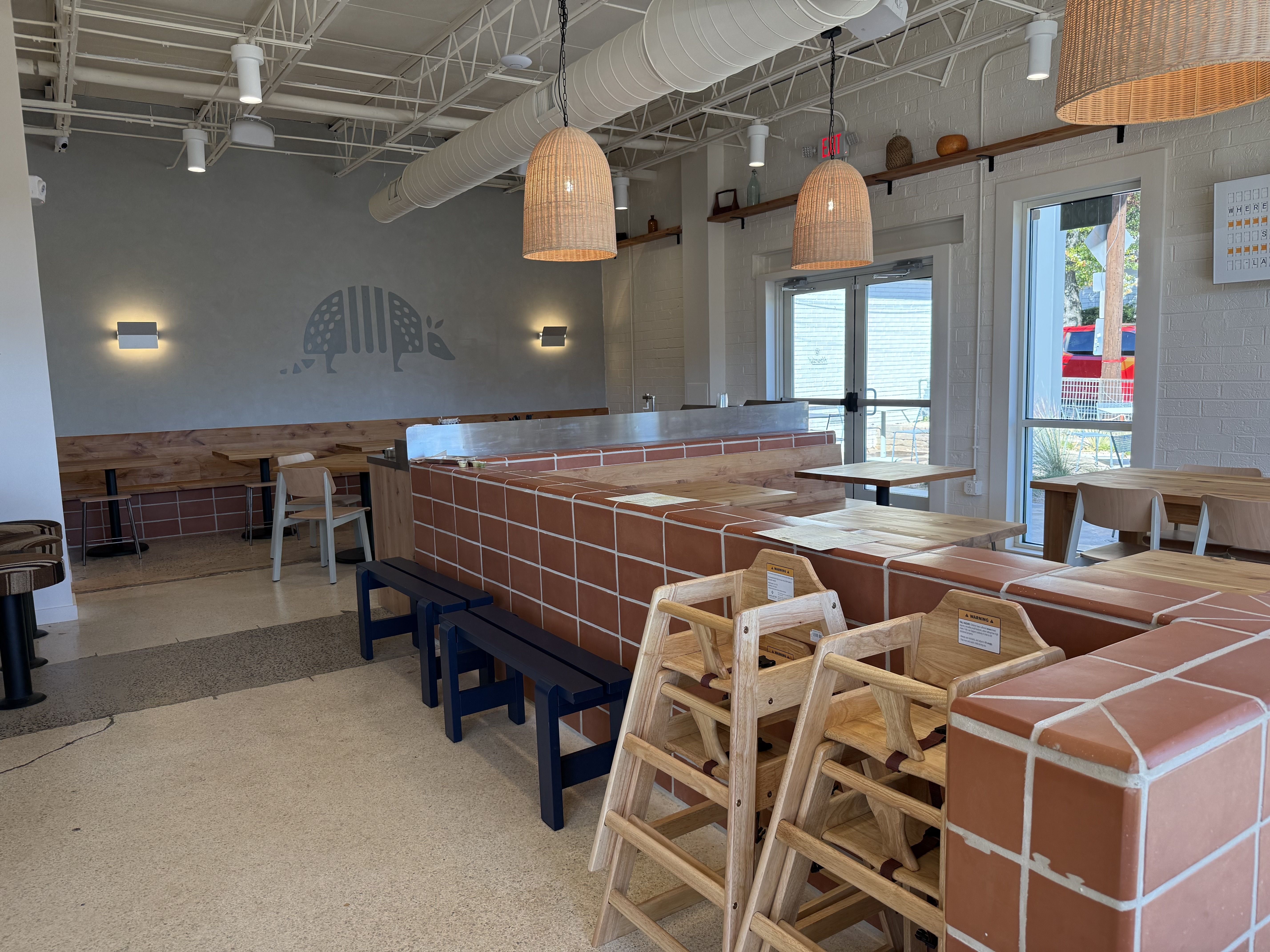 Modern cafe interior with white exposed ceiling, wicker pendant lamps, brick-tiled counters, and wooden tables. A gray wall shows a minimalist armadillo mural; glass doors.