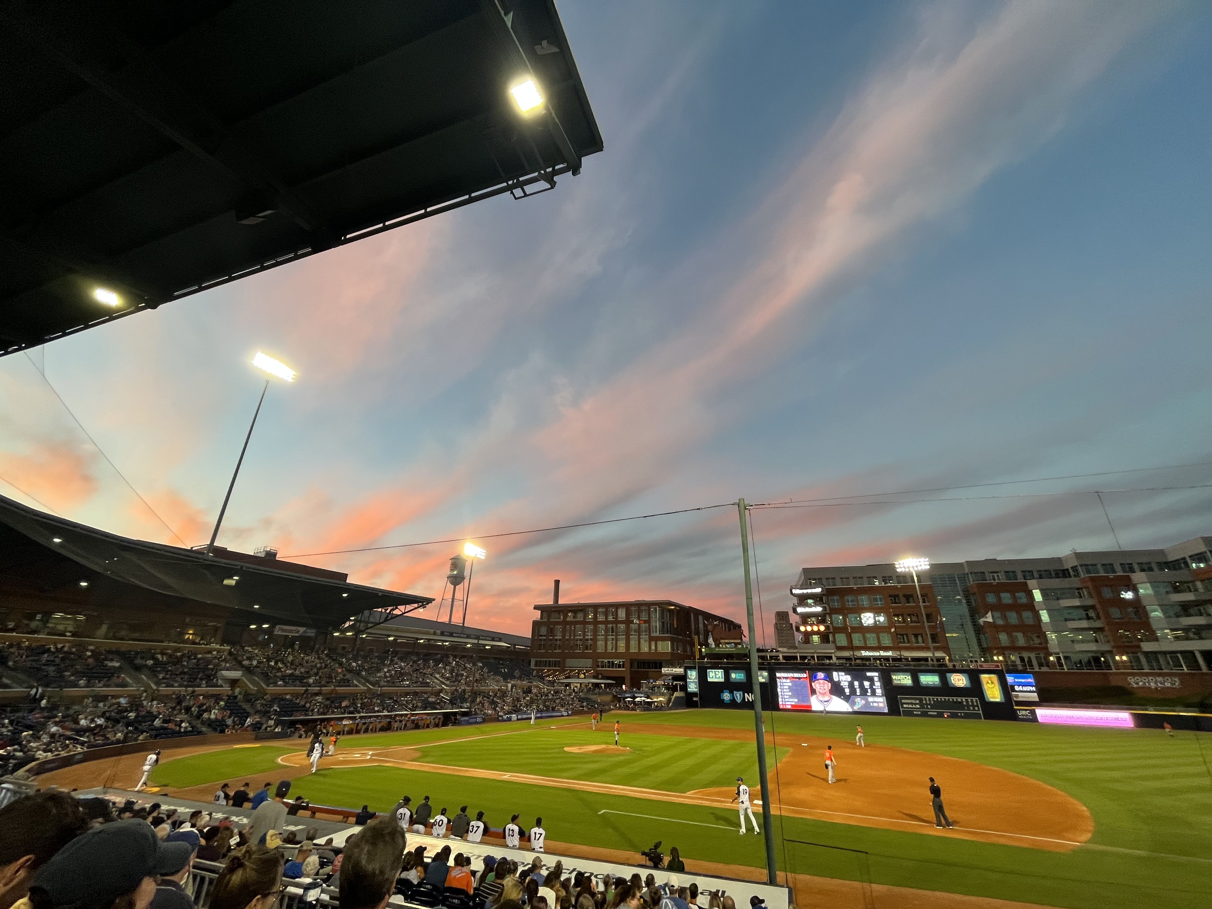 Baseball stadium at sunset, viewed from the stands beneath a dark overhang. Bright field lights glow as pink and blue clouds fill the sky; players occupy the green infield while spectators fill the seats.