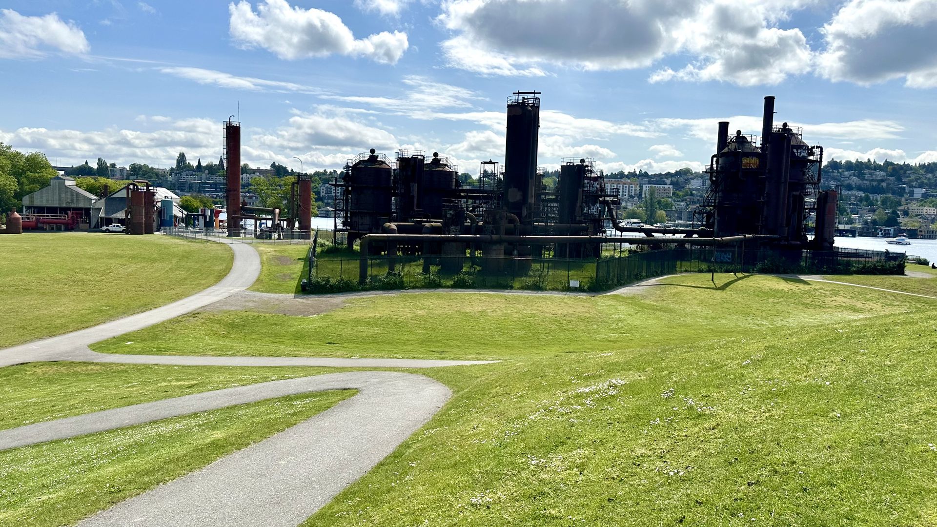 Gas Works Park is shown with the old refinery in the center and winding paths and grass in the foreground. Blue sky, clouds and the other shore across Lake Union can be seen in the background.