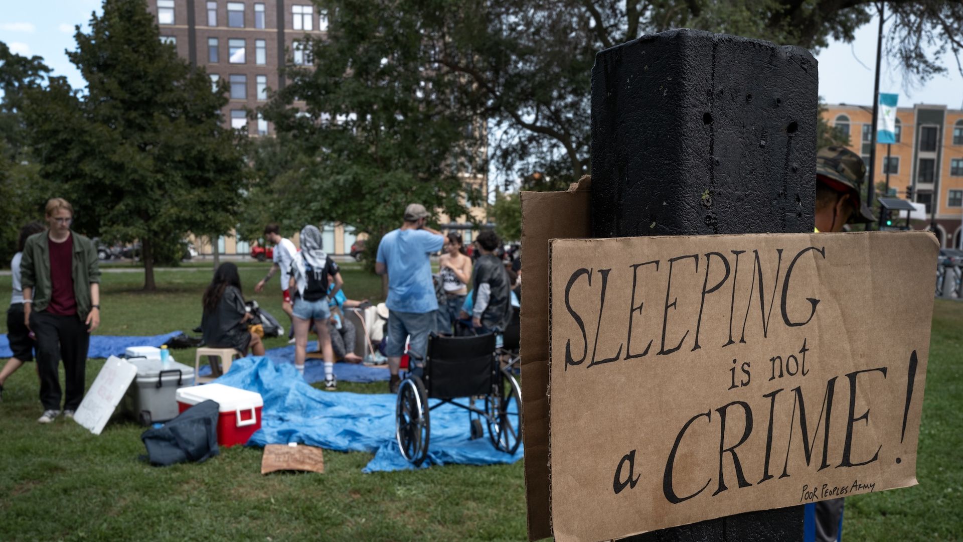 A brown paper sign that reads "Sleeping is not a crime" with people in the background in a park standing on top of a blue tarp.