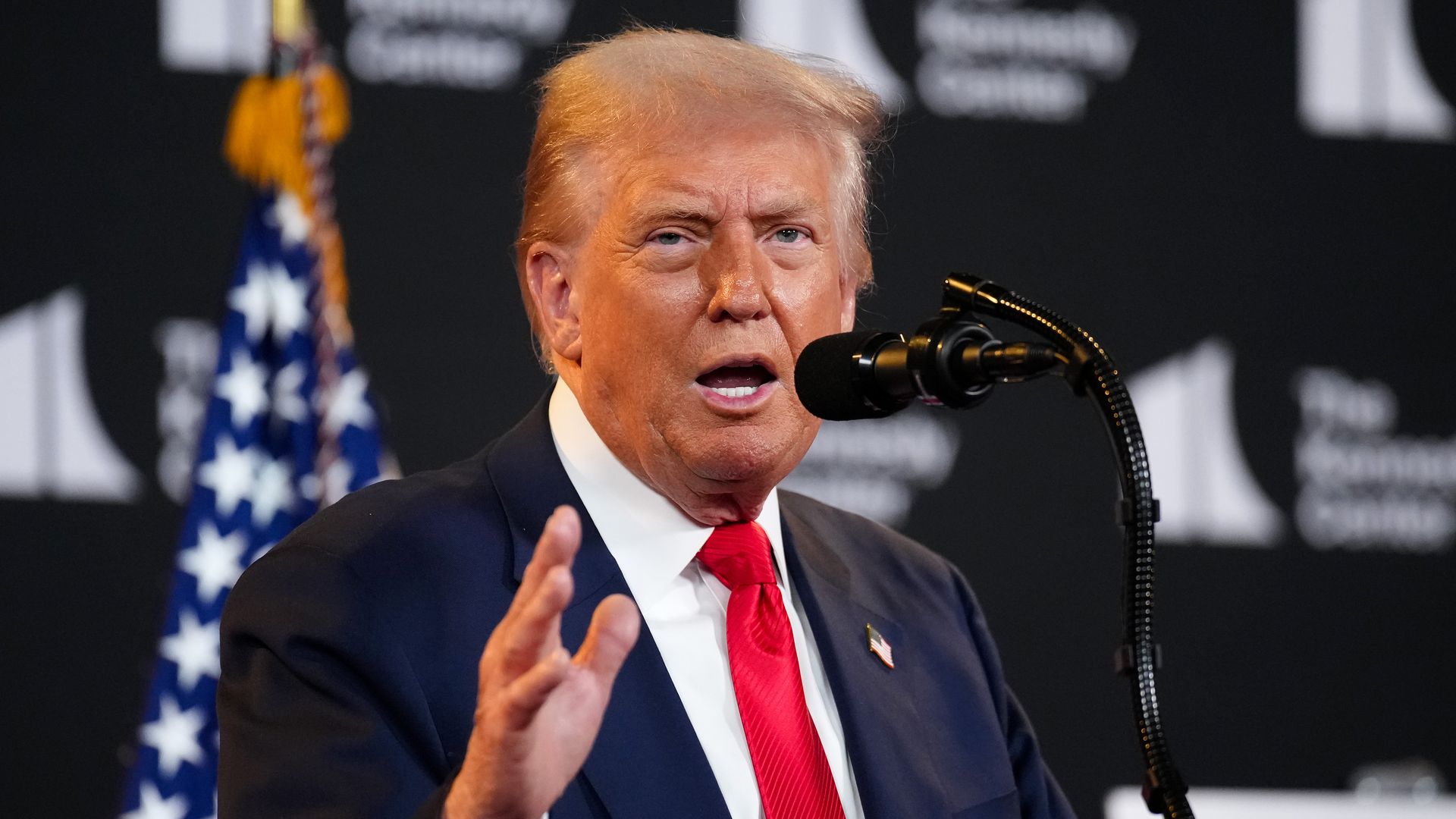 Man with orange-toned skin and blond hair wearing a dark suit, white shirt, and red tie speaking into a microphone with an American flag and black backdrop behind him.