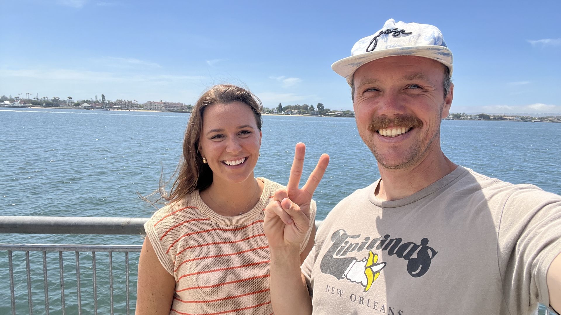Two people stand in front of water, one holding up the peace sign