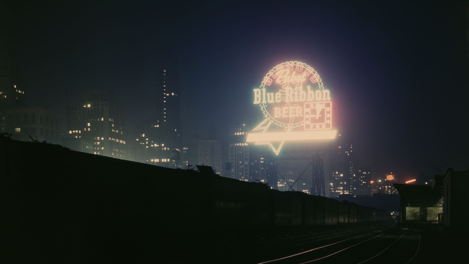 Night cityscape at dusk with a glowing neon sign reading "Blue Ribbon Beer" in a circular frame atop a structure near train tracks; dark buildings with lit windows fade into a hazy, foggy sky.