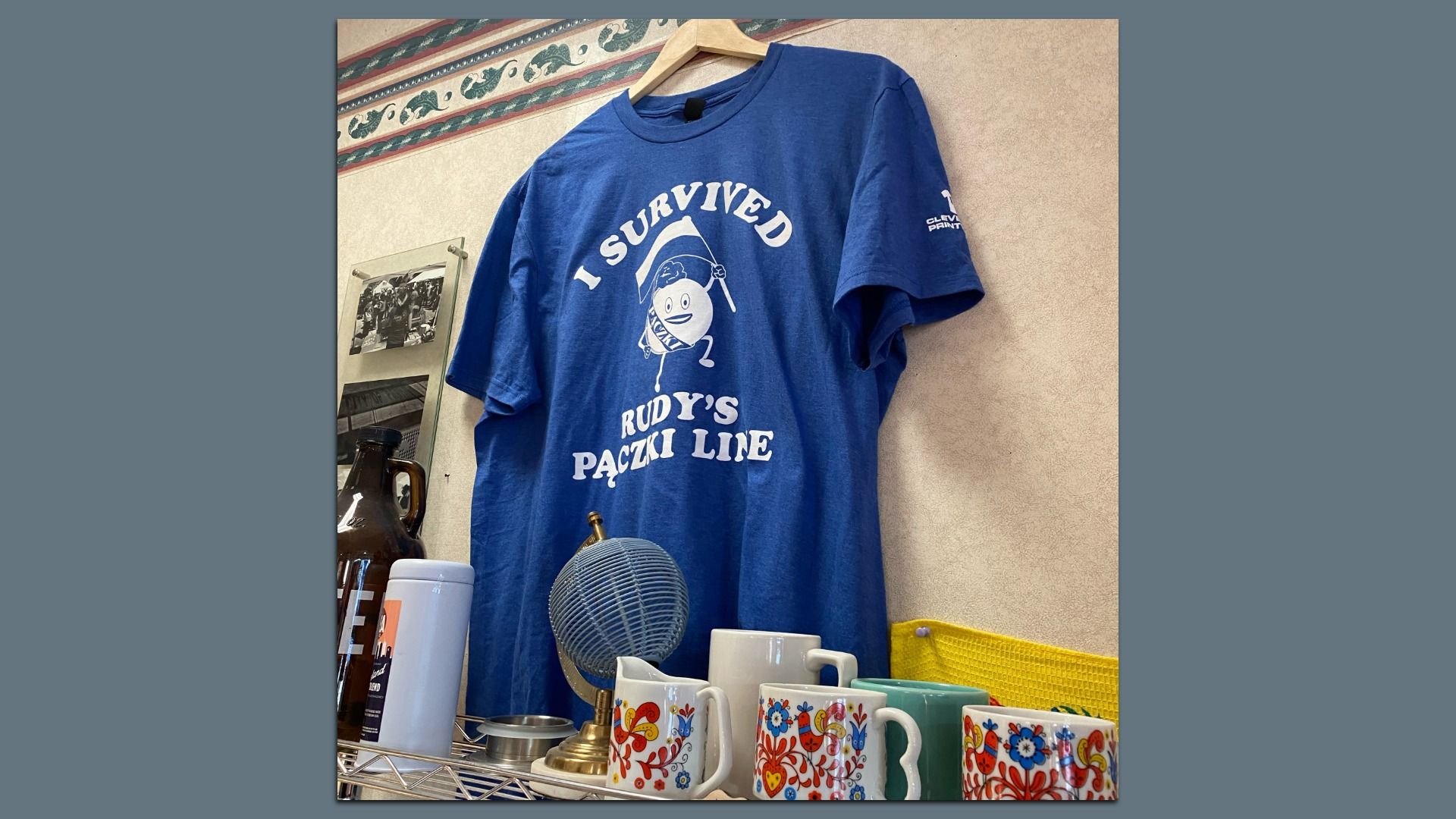 Blue T-shirt hanging on wall with white text and cartoon: "I Survived Rudy's Pączki Line." Below, colorful floral patterned mugs, a globe, and bottles on a wire shelf.