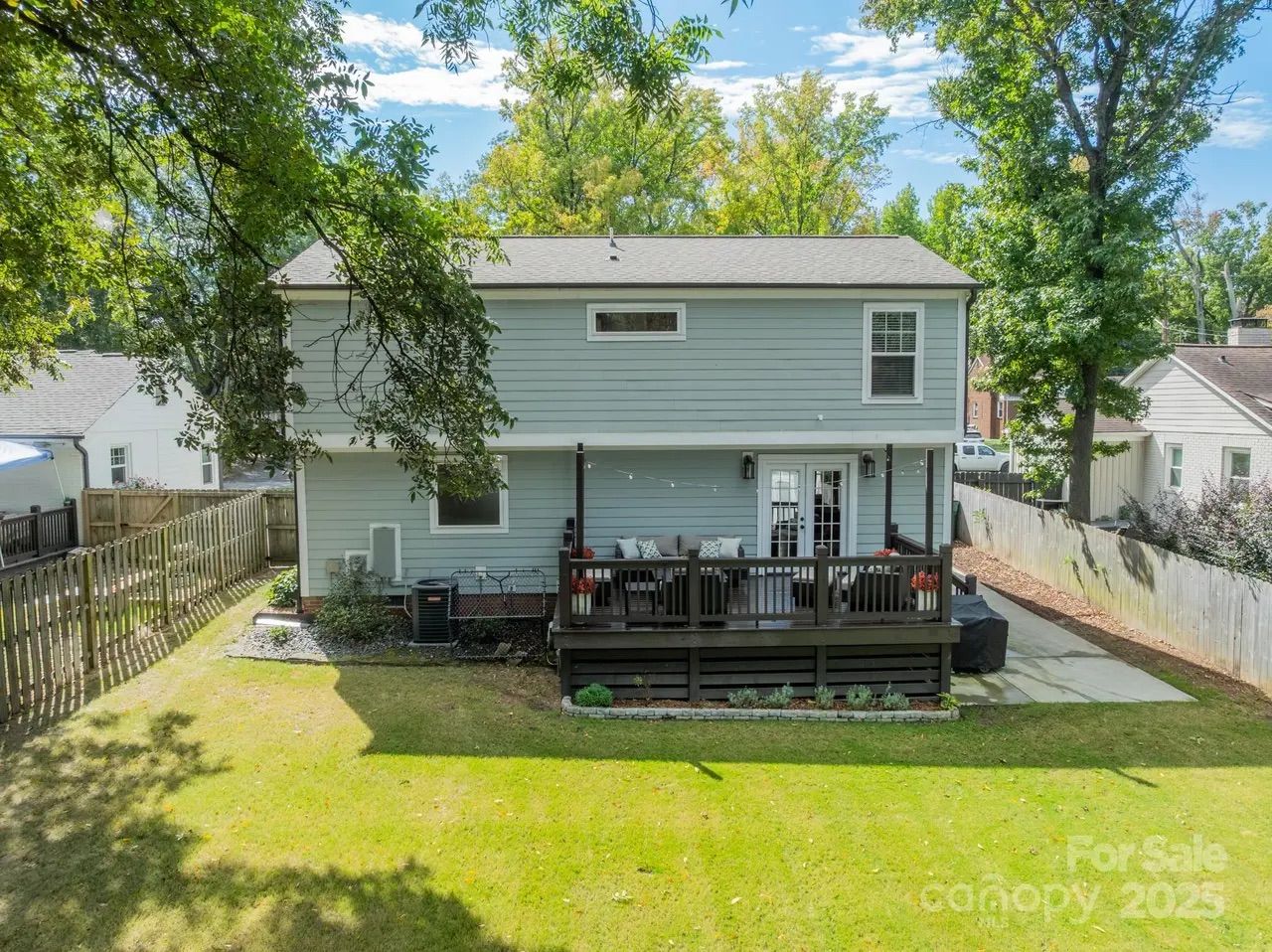 Two-story light blue house with a wooden deck in the backyard, surrounded by green lawn and tall trees under a partly cloudy sky.