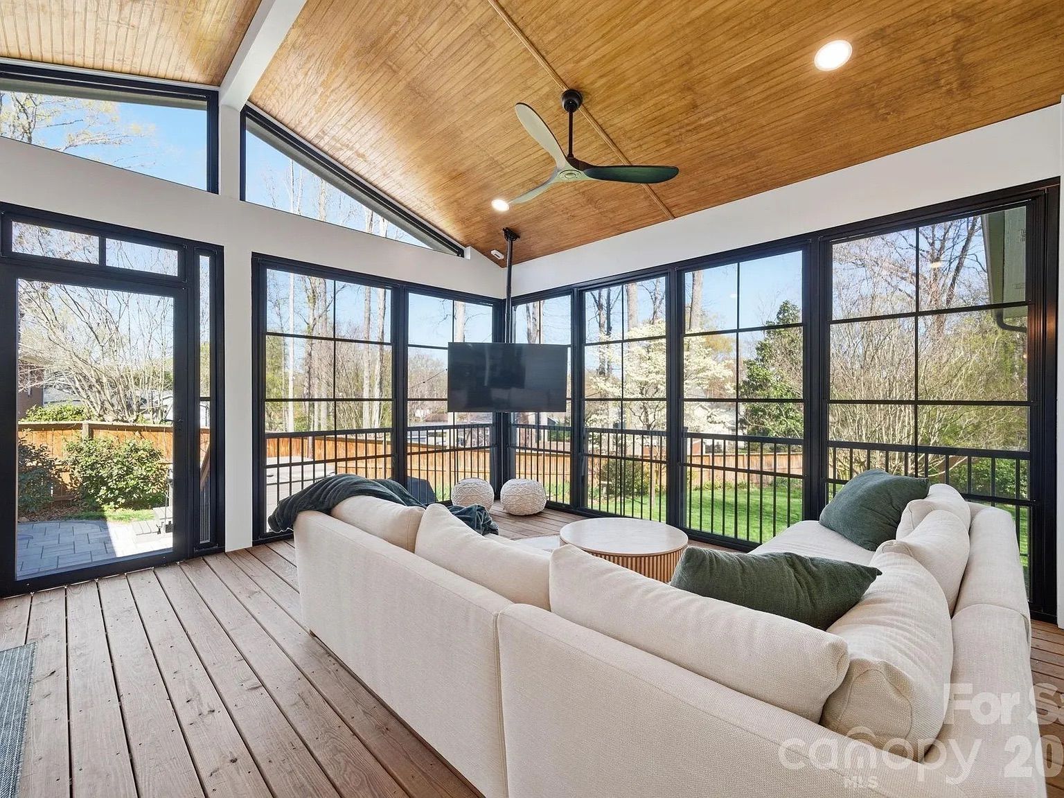 Bright sunroom with a warm wooden ceiling and black-framed glass walls. White sectional sofa with green pillows surrounds a round coffee table; a TV sits on a stand, and a fenced backyard is visible outside.