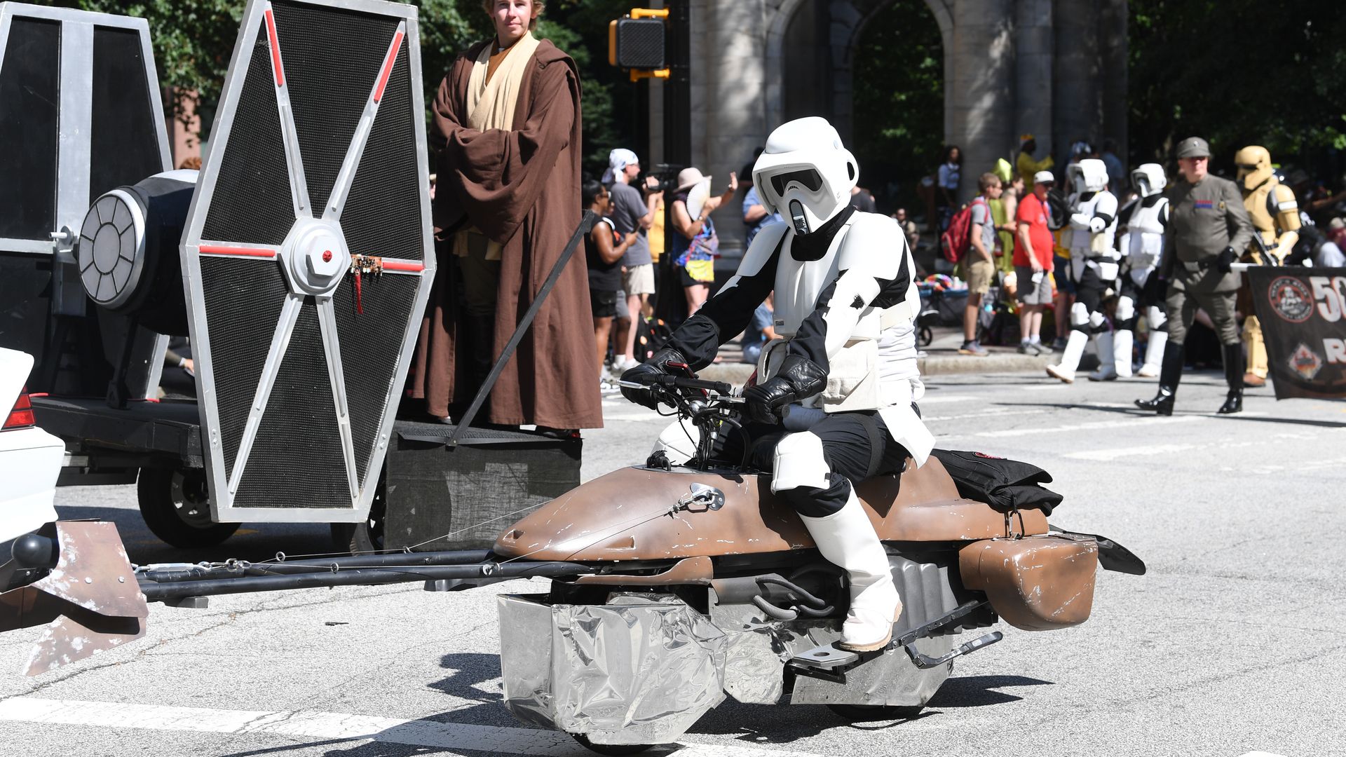 Person in white Star Wars scout trooper armor rides a brown speeder bike replica on a street during a parade, while another person dressed as a Jedi stands near a TIE fighter model.