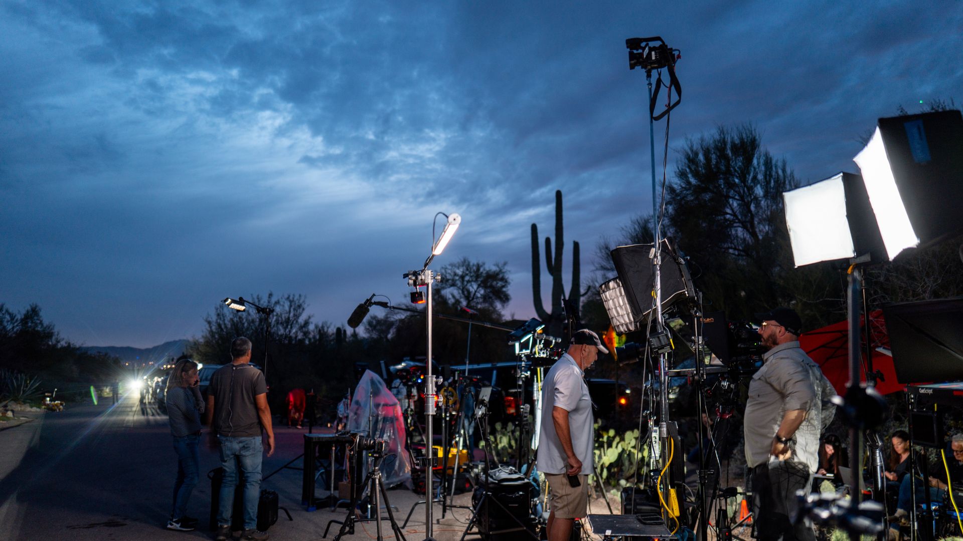 Film crew sets up cameras, lights, and microphones on a desert road at dusk, with cacti and a dark cloudy sky in the background. Several crew members are working and preparing equipment.