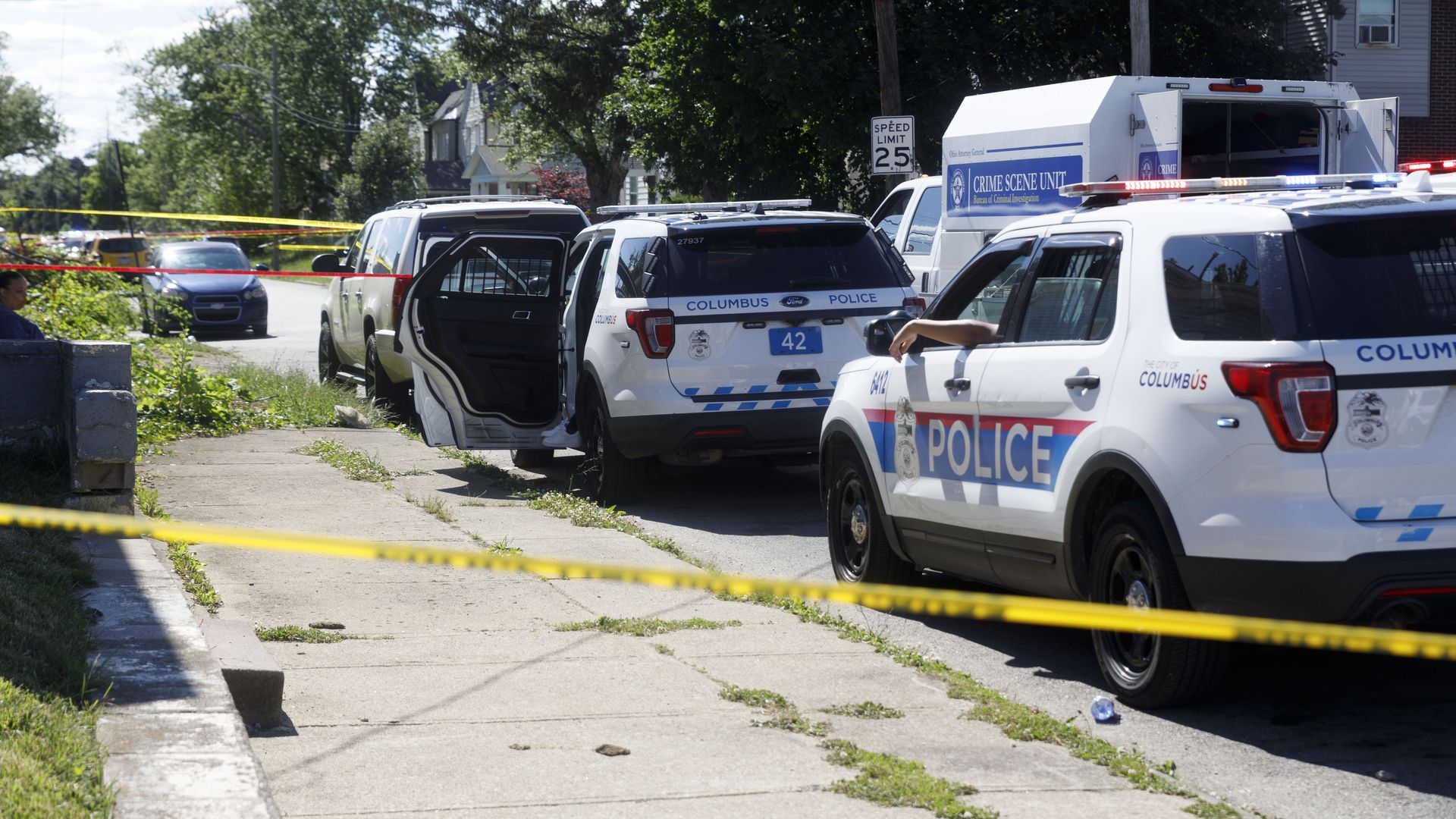 Law enforcement vehicles at the scene of a shooting, with crime tape in the foreground. 