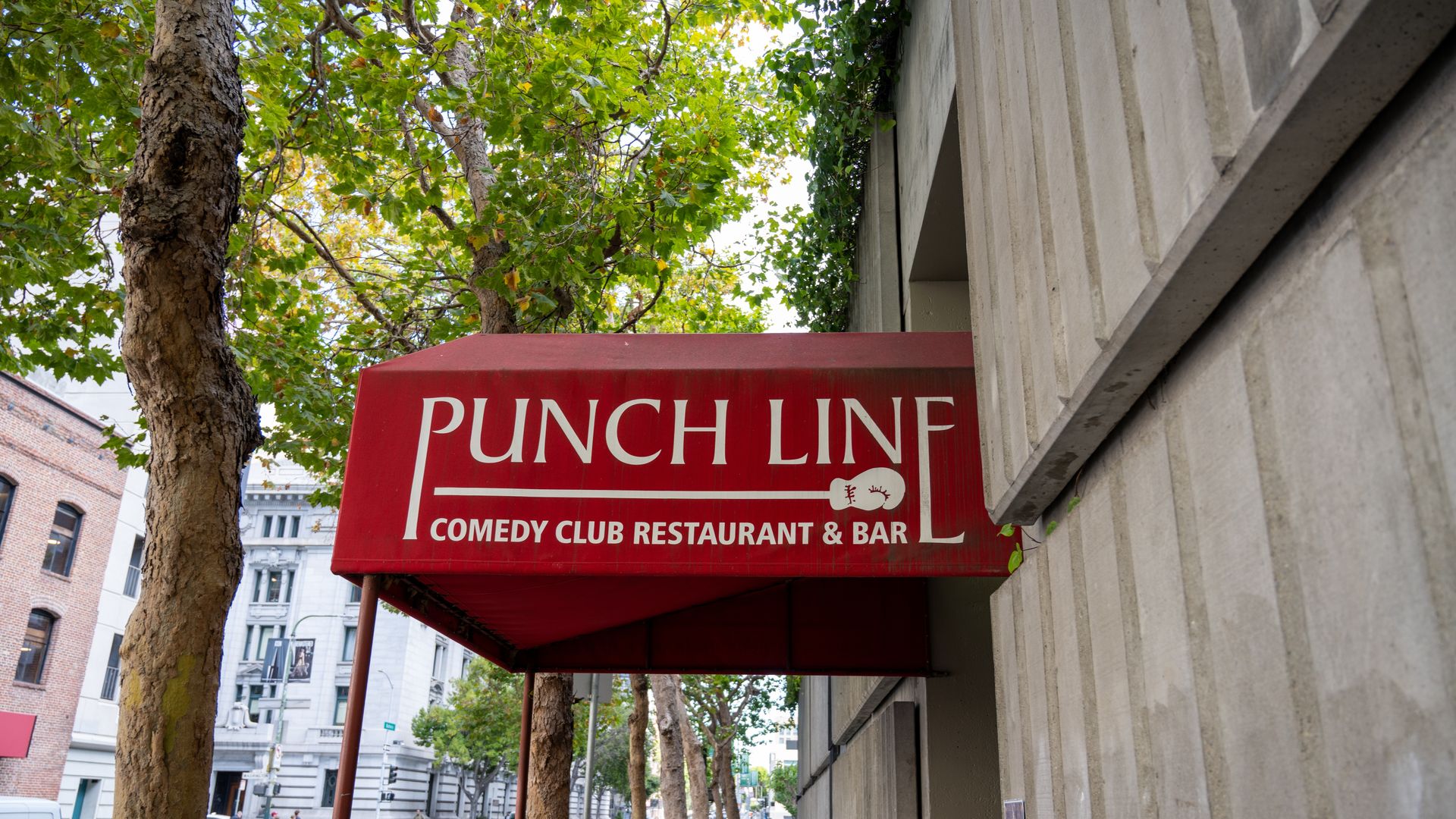 Red awning above a city sidewalk with white text reading "PUNCH LINE Comedy Club Restaurant & Bar." Trees and buildings are visible in the background.