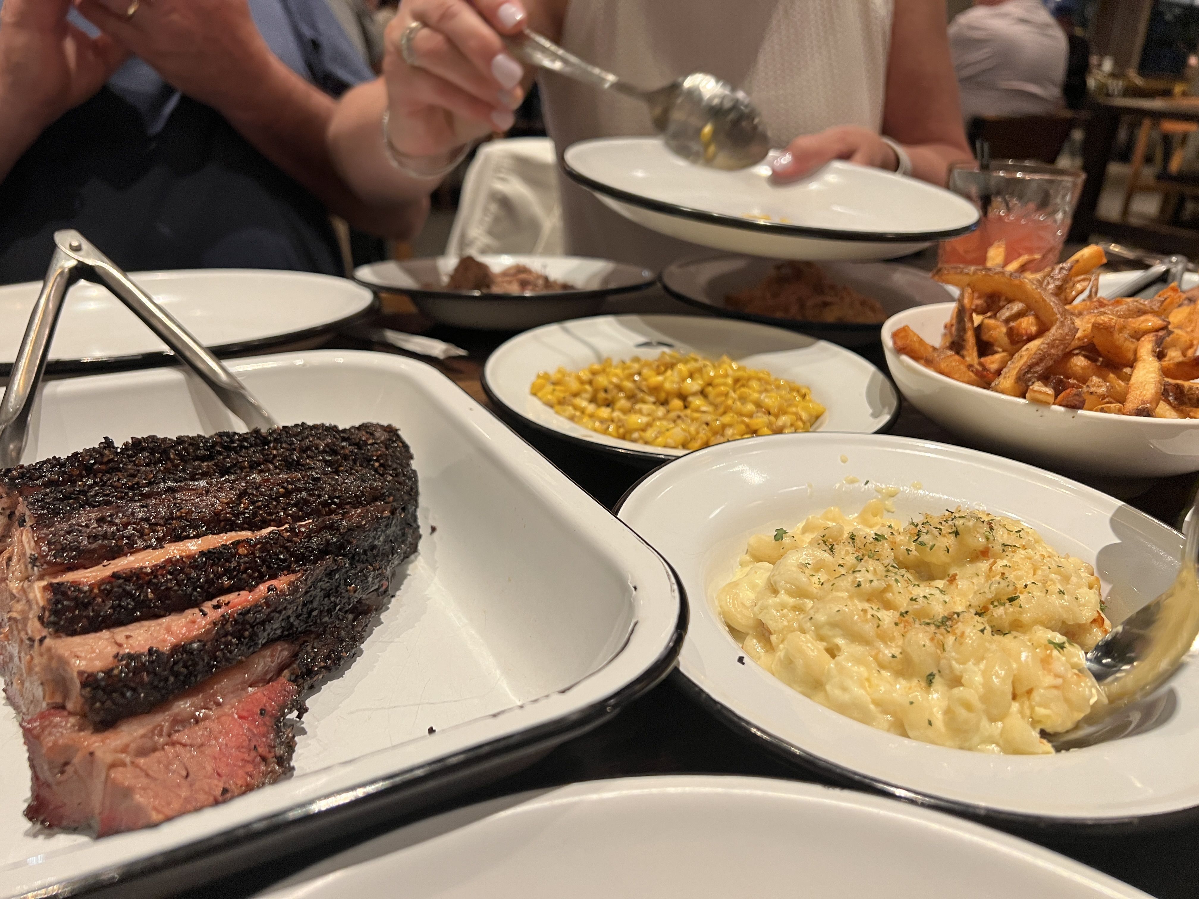 Close-up of a family dinner: a dark-crusted roast beef in the foreground with macaroni and cheese, corn, and roasted potatoes; hands serve onto plates at a shared table.