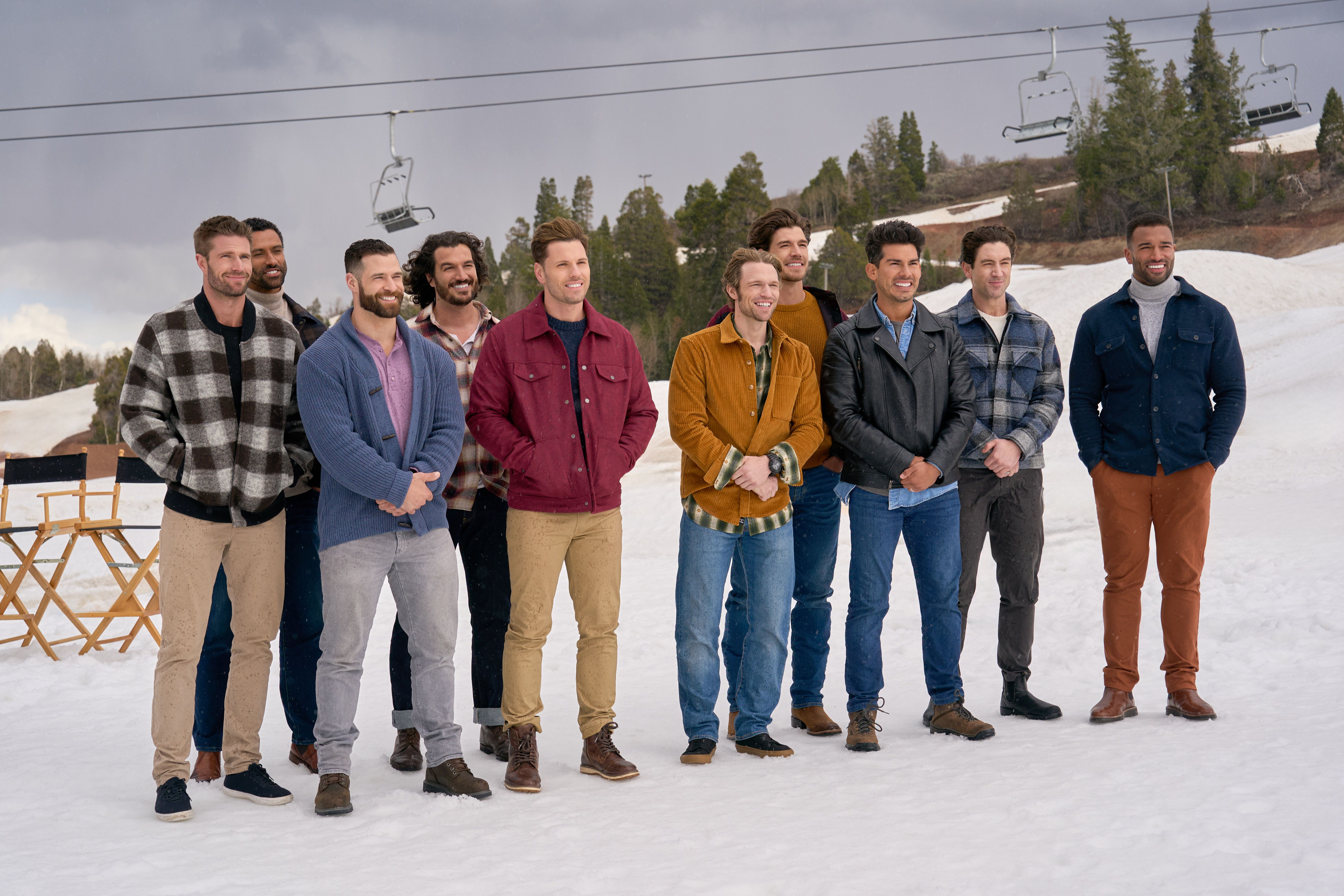 Ten men standing in snow wearing casual jackets and sweaters, smiling. Chair lift and pine trees in the snowy background under a cloudy sky.