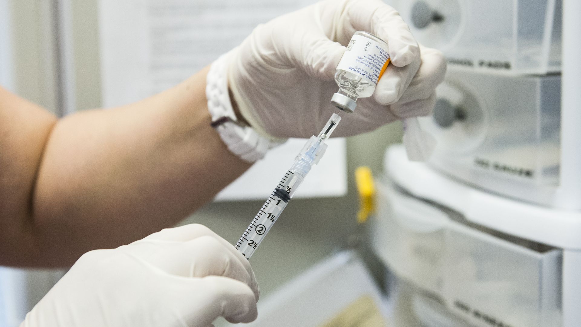 Gloved hand draws liquid from a small vial into a syringe for vaccination, with medical equipment blurred in the background.