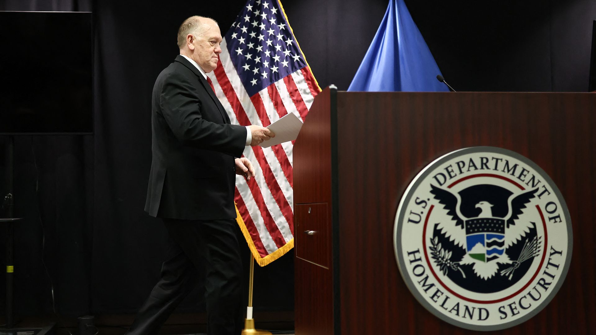 Man in black suit holding paper walking past podium with U.S. Department of Homeland Security seal, with American flag and blue flag in background on black stage.