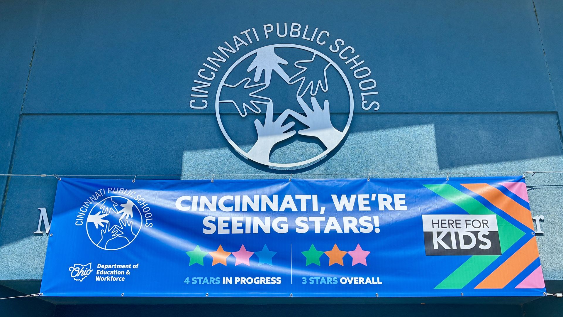 Blue wall with a large circular Cincinnati Public Schools logo featuring interlocking hands. A blue banner below reads "CINCINNATI, WE'RE SEEING STARS!"