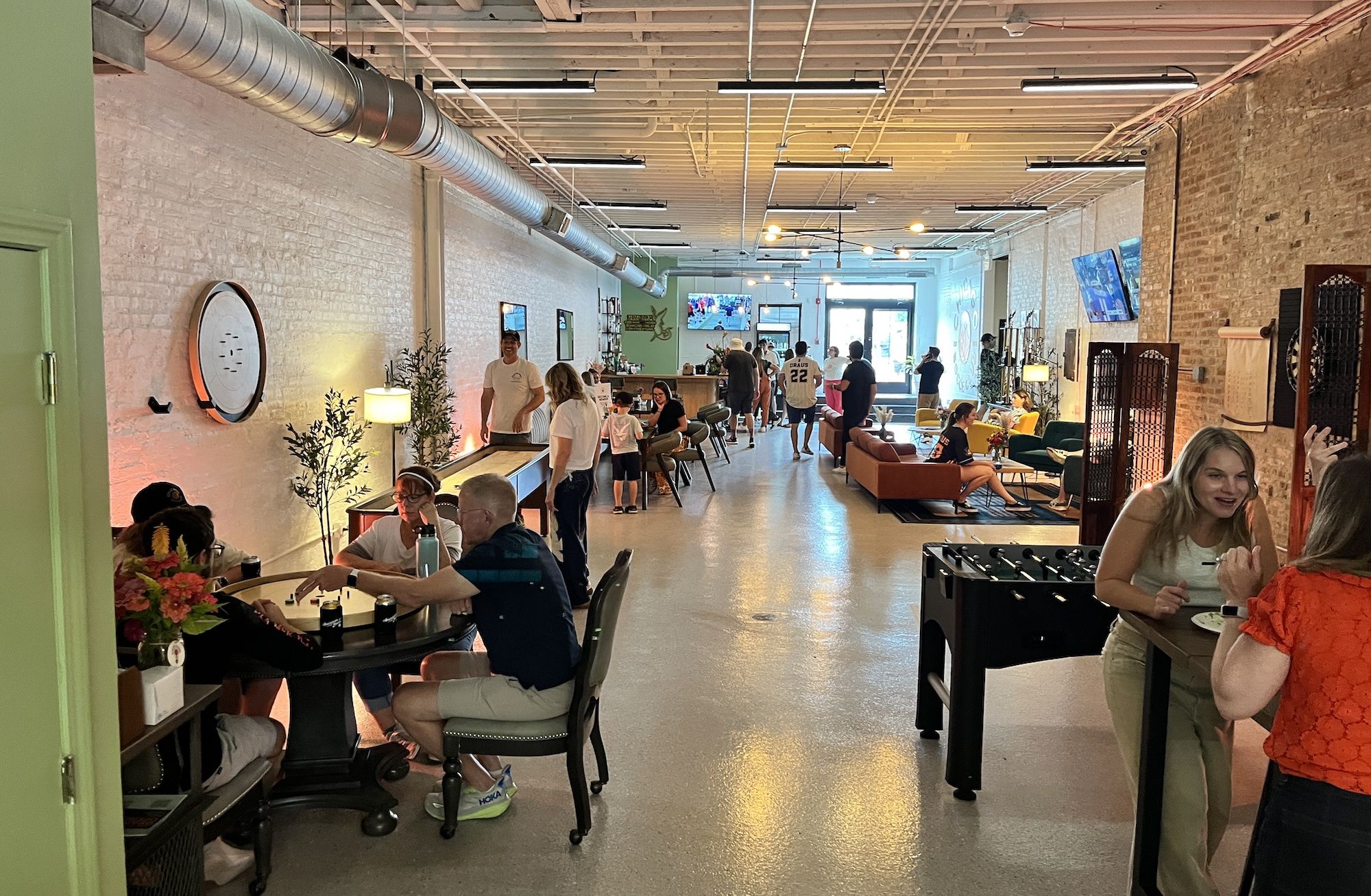 Industrial cafe with exposed ducts and white brick walls. Patrons sit at tables and colorful couches; a foosball table in the foreground, wall screens, and a bright back entrance.