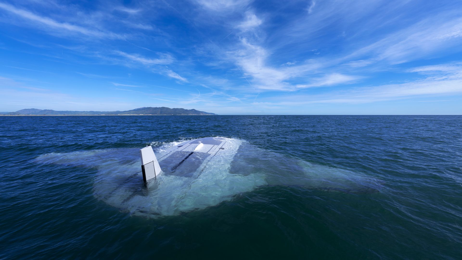 A massive almond-shaped underwater drone, grey in color, breaches blue waters in the foreground. California mountains and blue skies can be seen in the background.