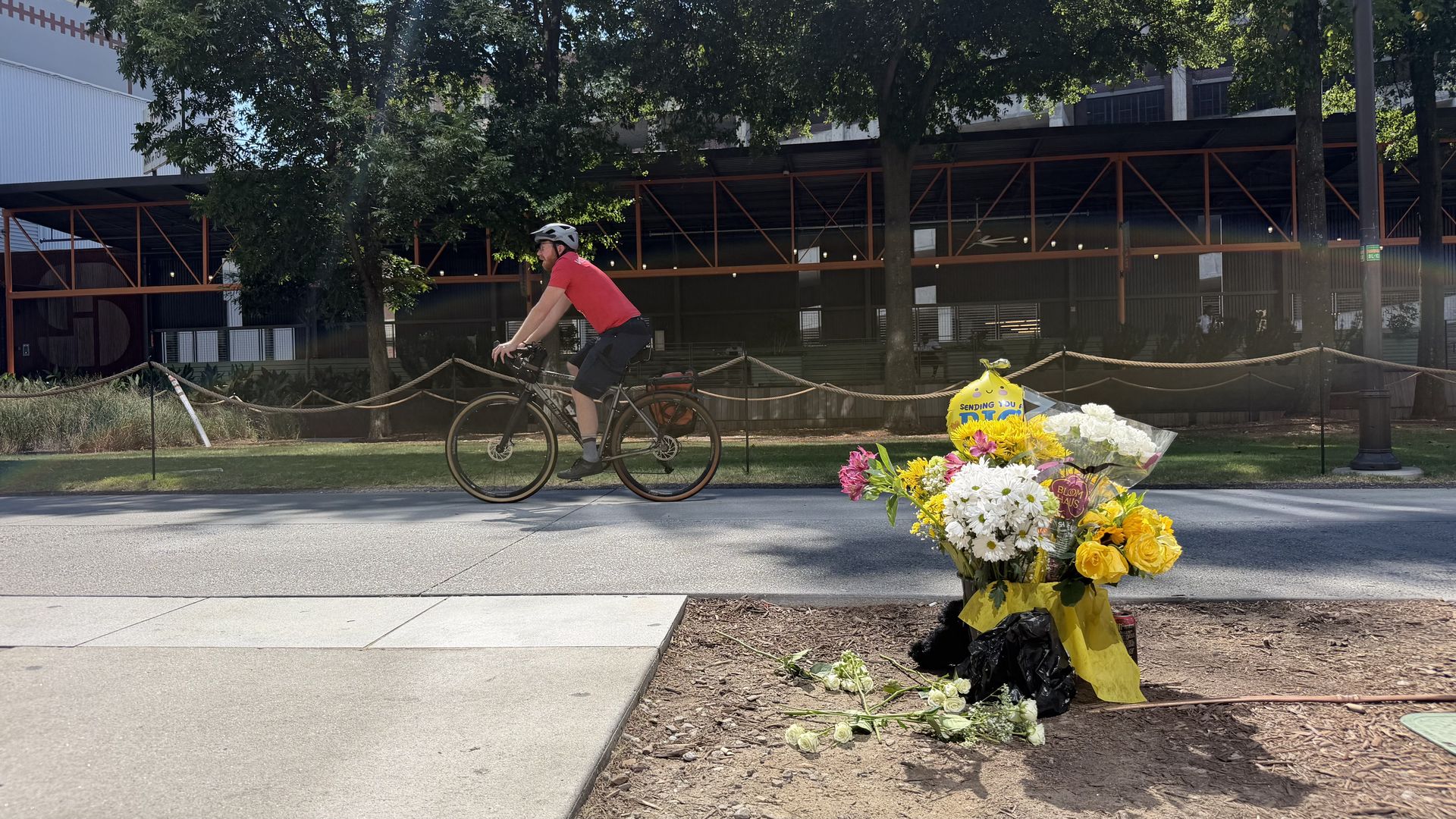 Man in a red shirt wearing a helmet rides a black bike along a sidewalk in front of a building with orange framework; a bouquet of yellow and white flowers sits on the curb in the foreground.