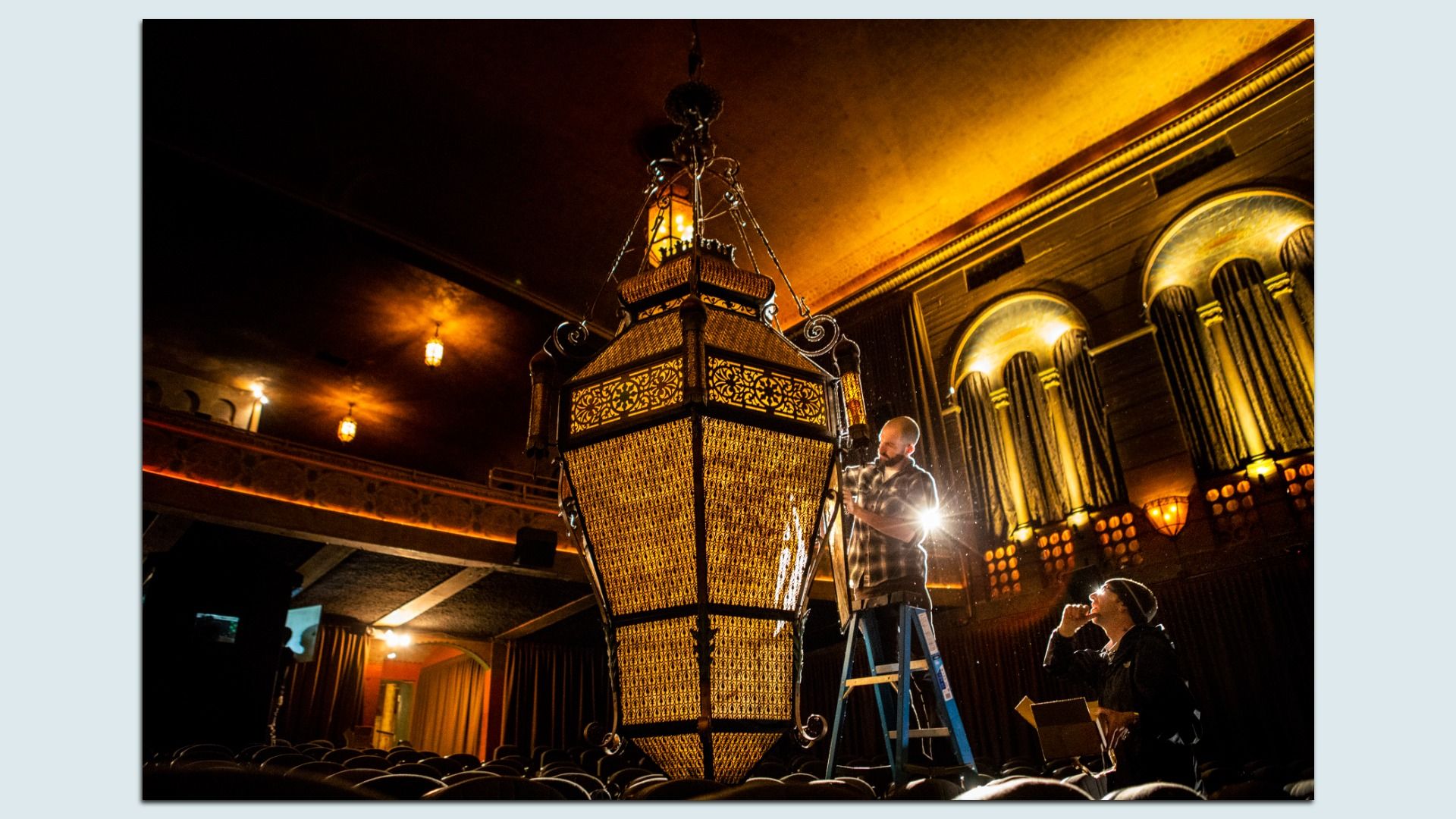 Inside a dimly lit theater, a large ornate chandelier hangs from the ceiling while a man on a ladder adjusts it. Warm amber lights illuminate arched columns and rows of seats below.