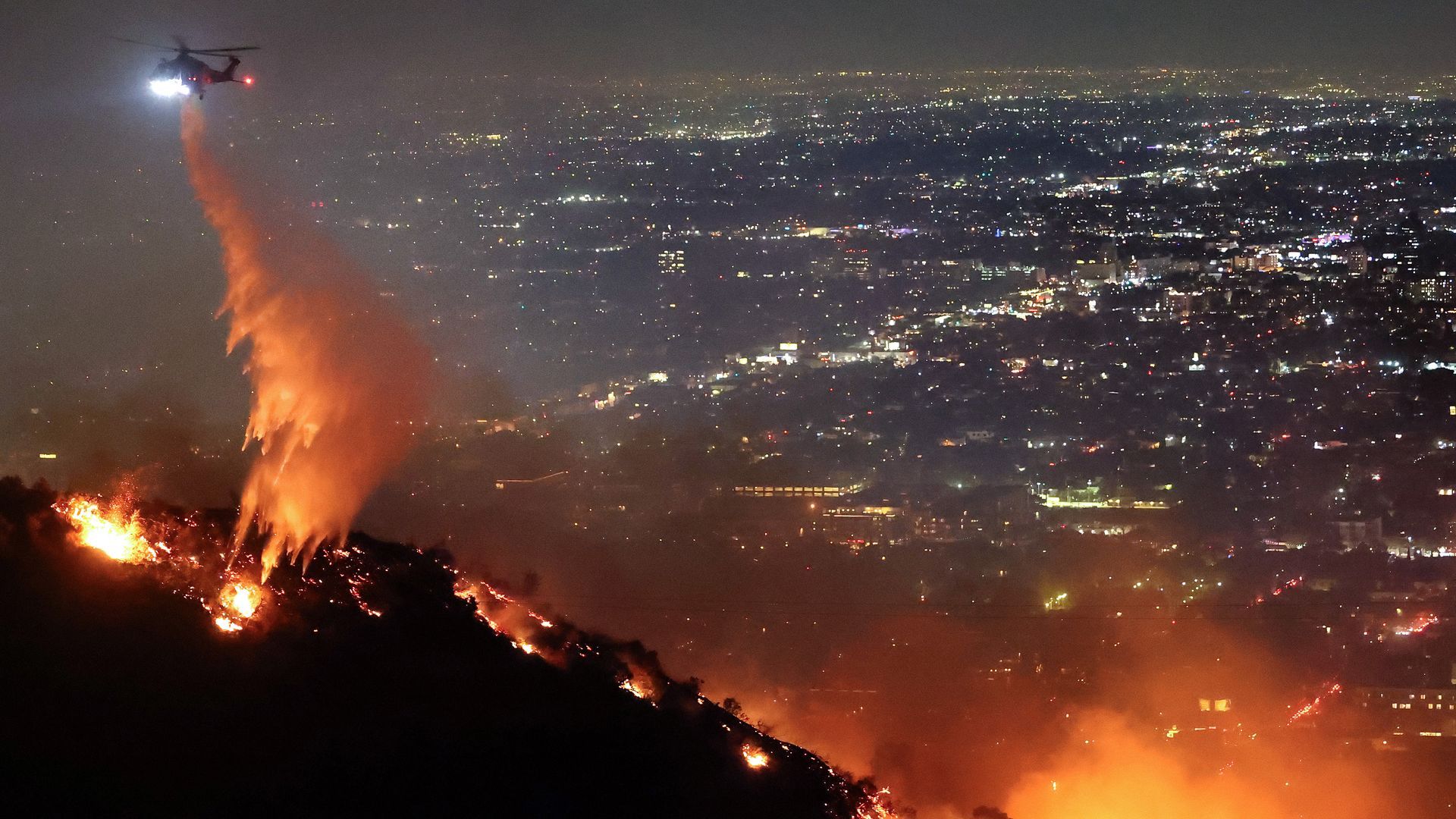 A helicopter drops water on a fire in the Hollywood Hills with the city lights in the distance.