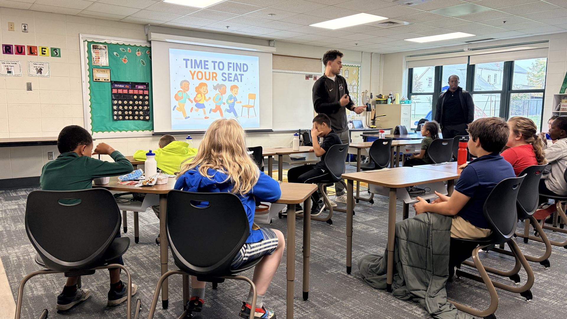 Bright classroom with students facing a projector showing the slide "TIME TO FIND YOUR SEAT" with running children. A teacher in a dark jacket speaks at the front; another adult stands by windows.