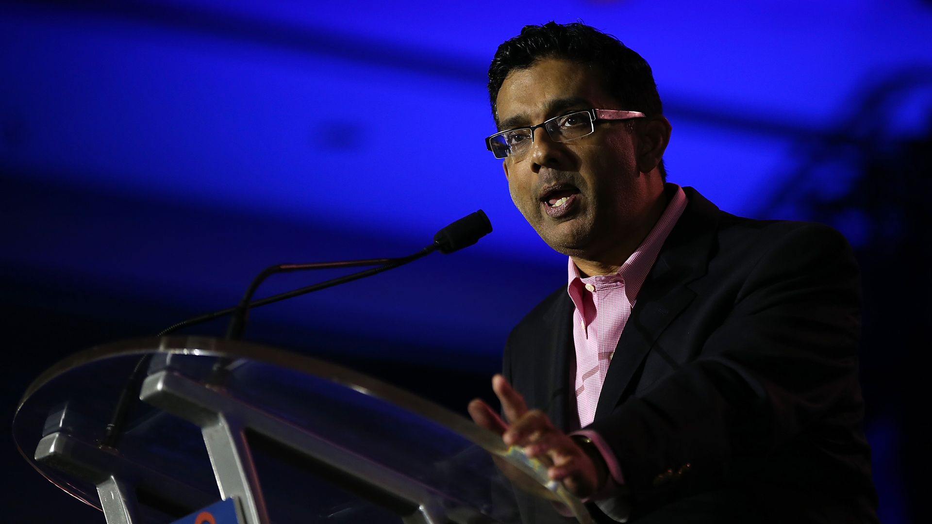 Dinesh D'Souza speaking at a podium before a blue background.