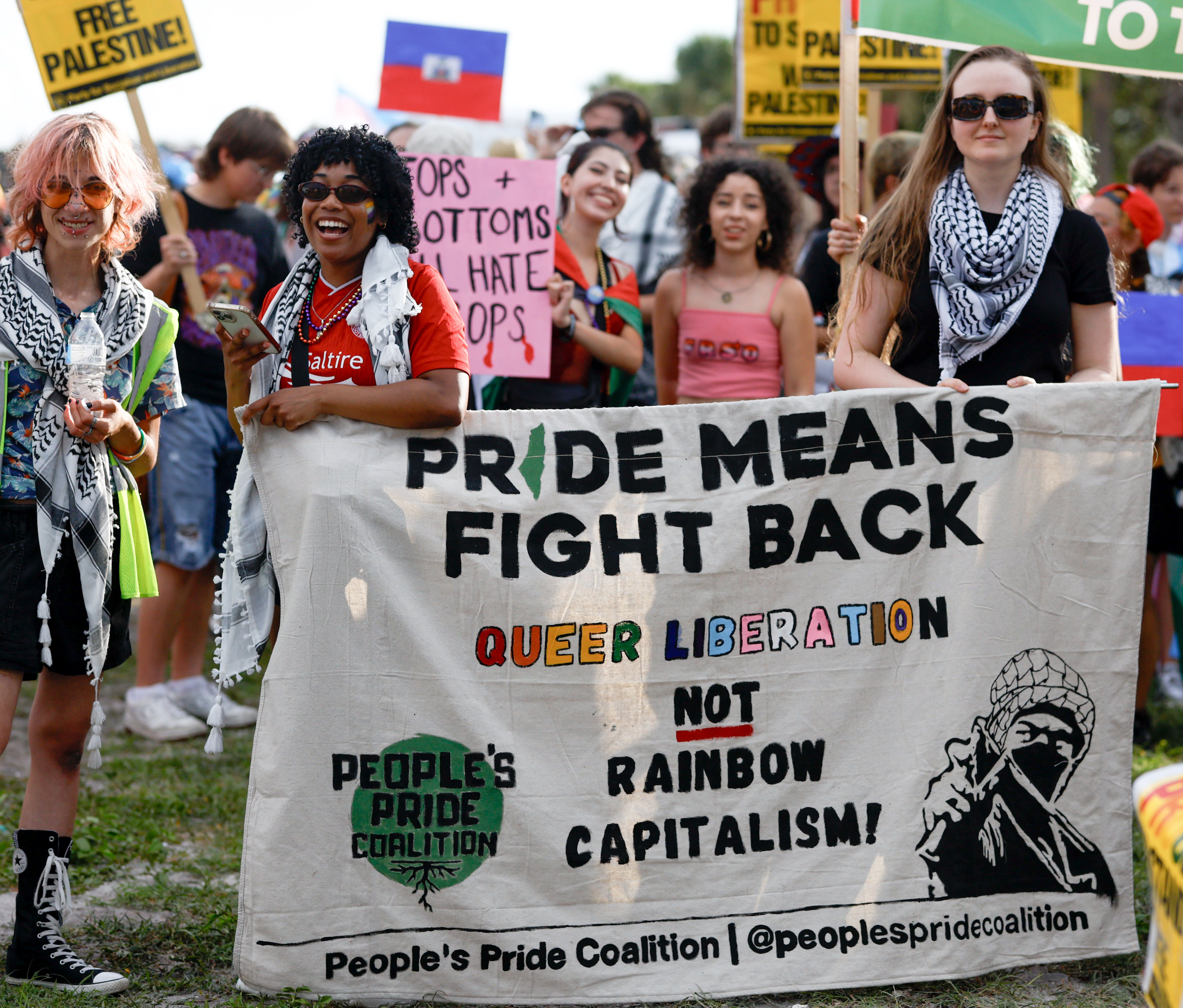 A group of people wearing keffiyehs hold a sign that reads, "PRIDE MEANS FIGHT BACK, QUEER LIBERATION NOT RAINBOW CAPITALISM." A green logo on the bottom lefthand side of the sign reads, "People's Pride Coalition." Black text on the bottom reads, "People's Pride Coalition | @peoplespridecoalition."
