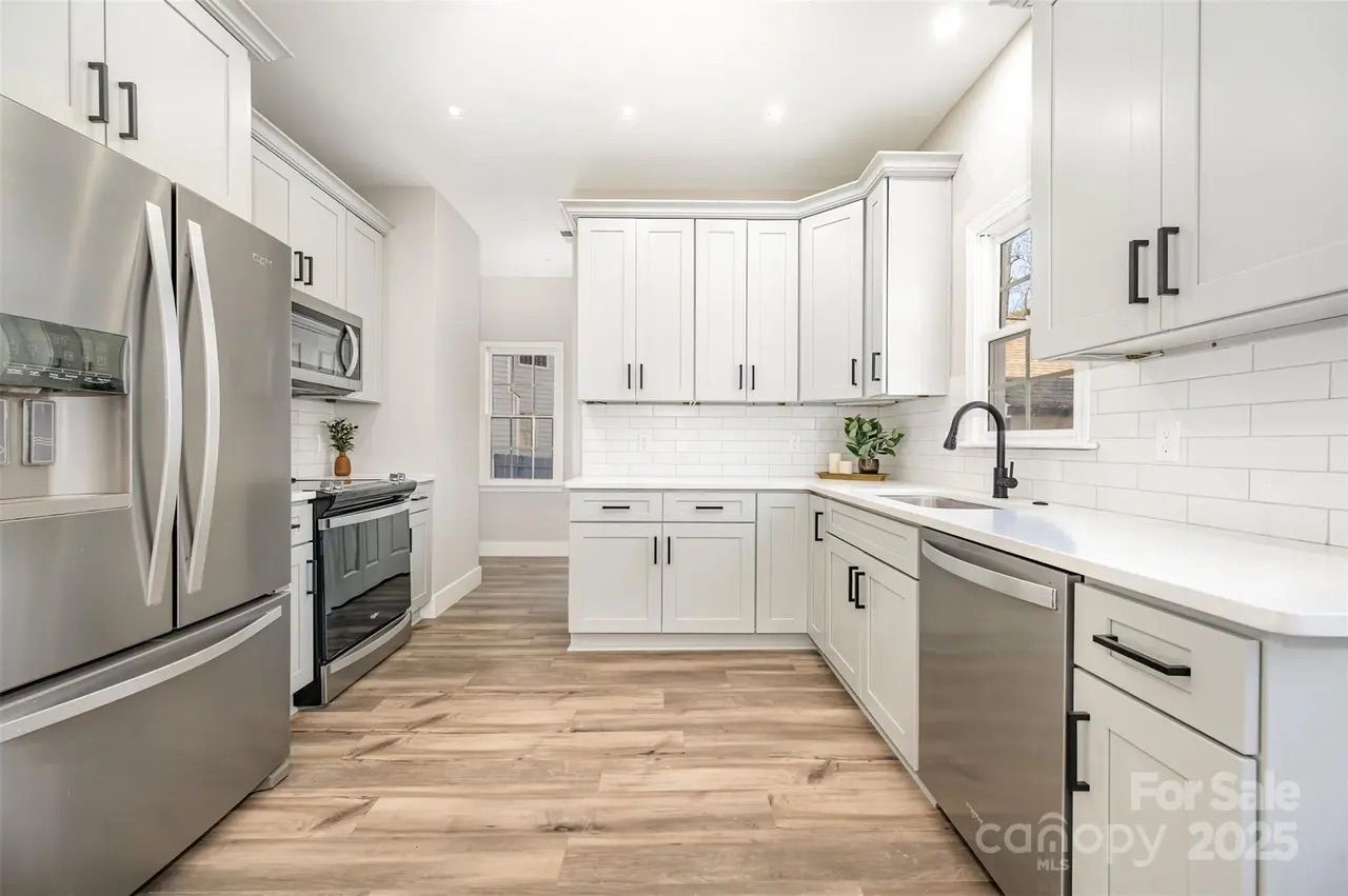 Modern kitchen with white cabinets, stainless steel refrigerator, oven, microwave, dishwasher, black handles, wood-look flooring, white countertop, subway tile backsplash, and small green plants.