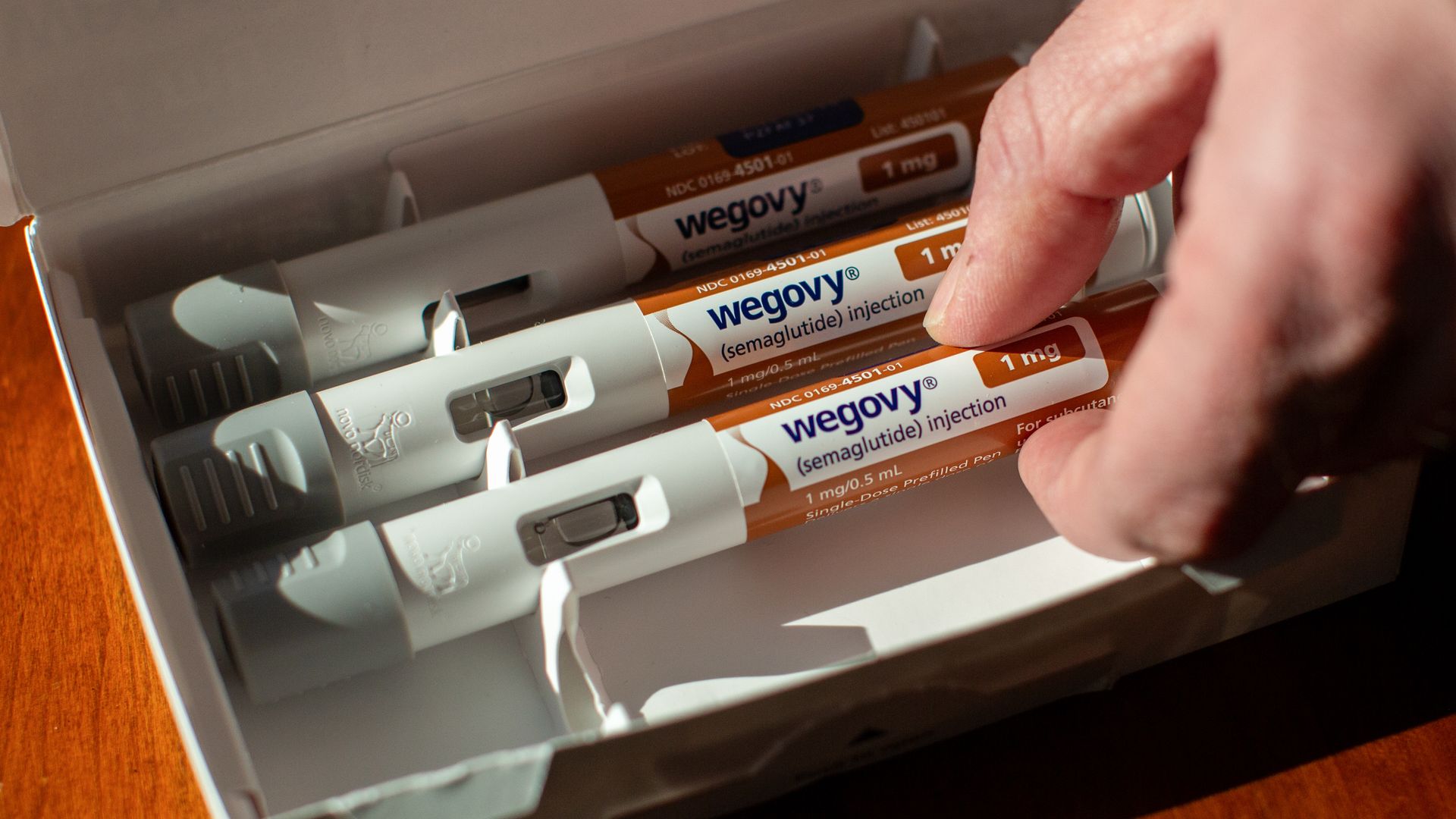 Hand picking a Wegovy (semaglutide) 1 mg injection pen from a box containing three pens on a wooden surface.