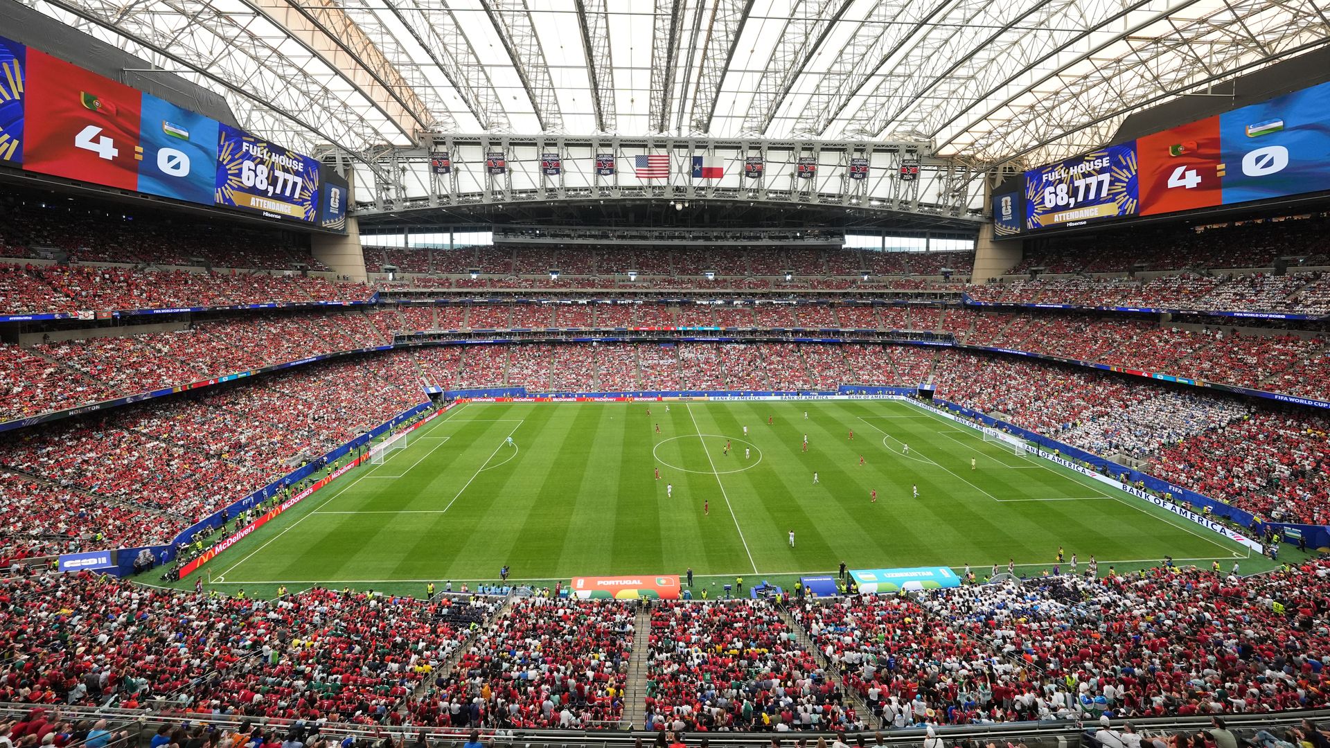 Interior of a large soccer stadium filled with red-clad fans. A sweeping glass roof, colorful banners and screens hanging over the stands show a 4-0 score and 68,777 attendance.