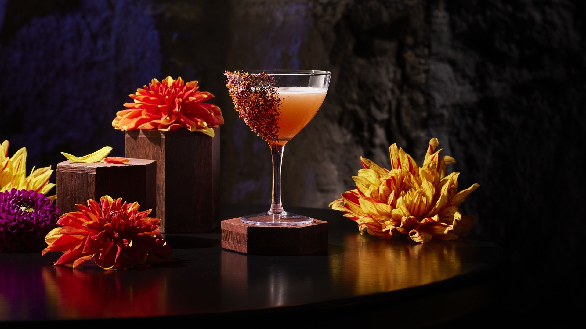 An orange cocktail with tajin on the side in a coup glass, next to colorful flowers.