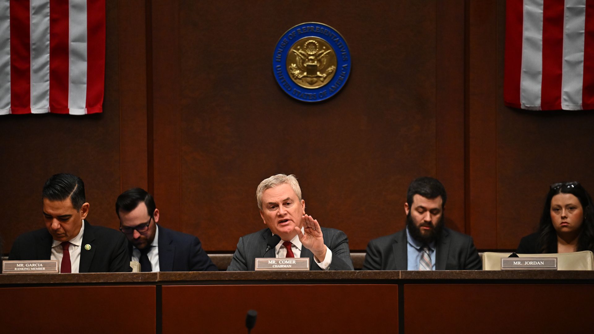 Congressional hearing panel at a wood desk; center speaker Mr. Comer raises his hand while speaking. U.S. flags flank the stage and the House seal hangs overhead.