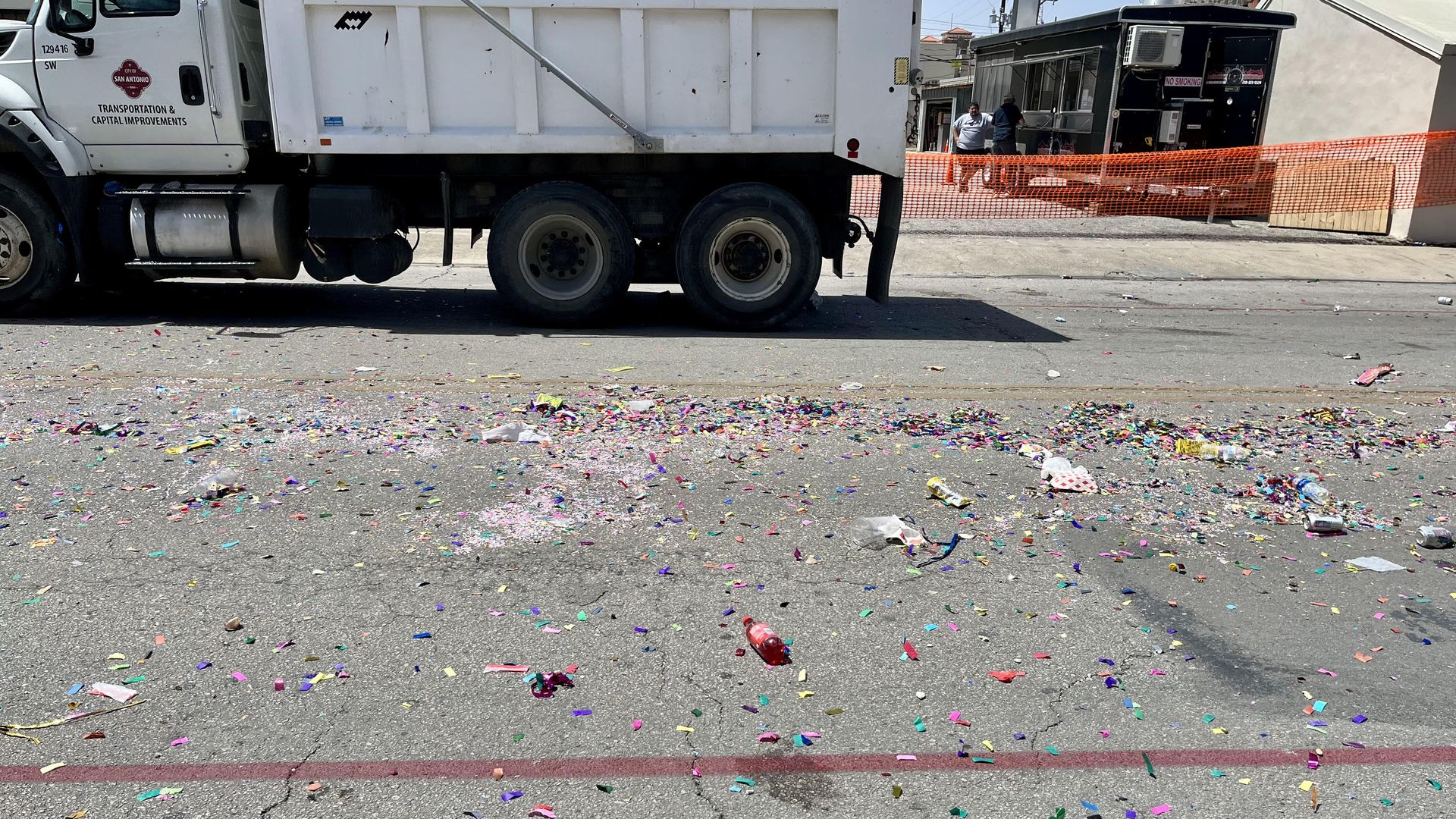 A street is left covered in colored confetti and a plastic bottle of Big Red. In the background is a city garbage truck passing by.