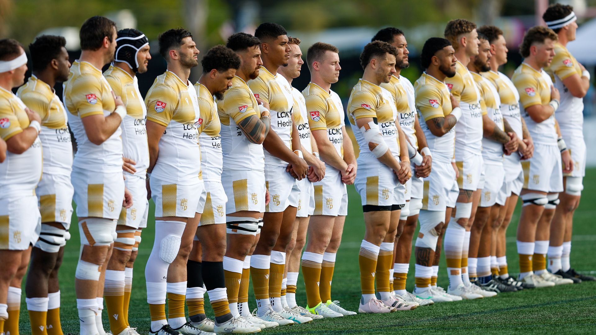 Rugby team in white and gold uniforms lined up on a green field, standing with heads bowed, some with hands over hearts, preparing for a game or anthem.