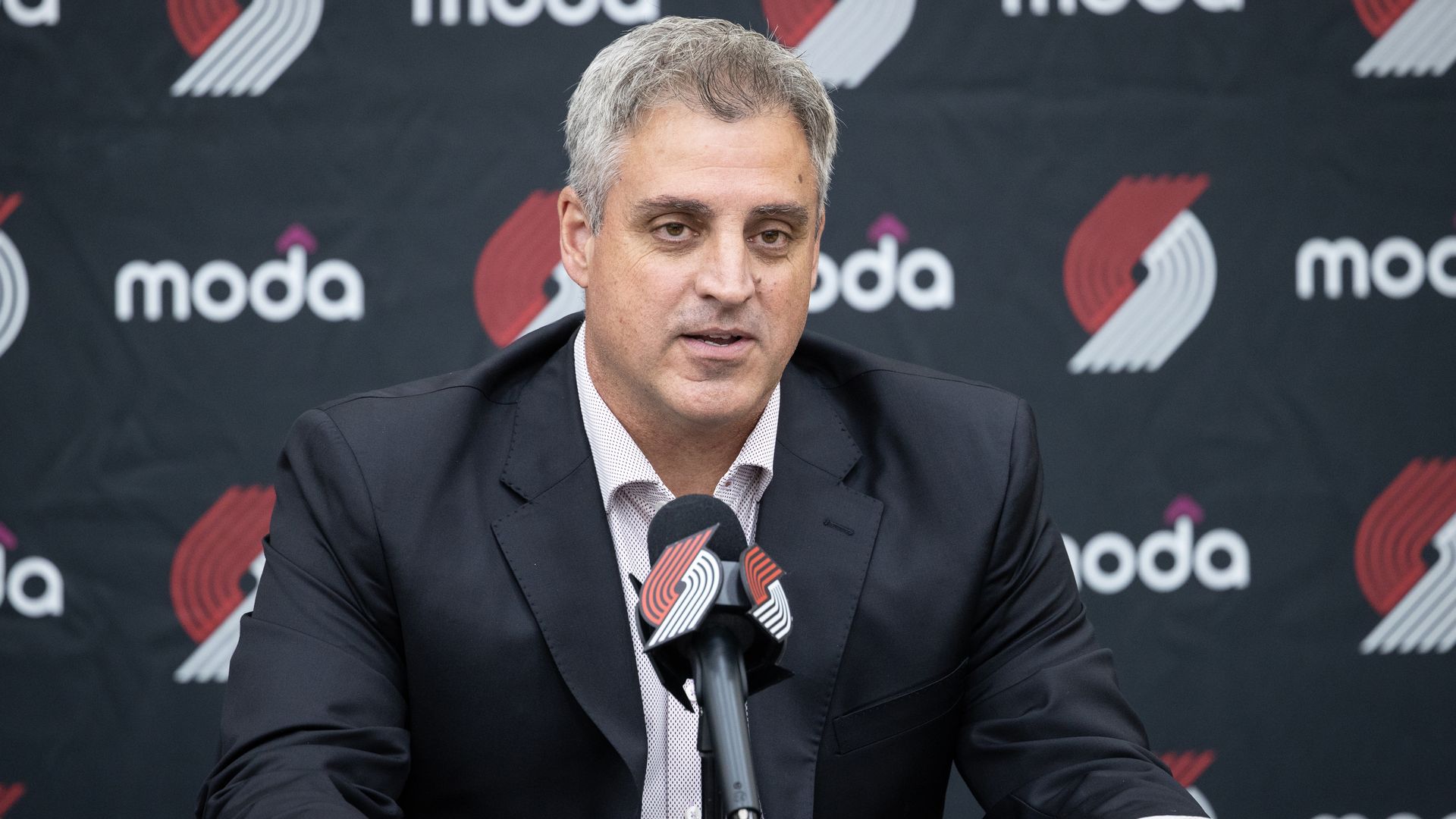 A man with gray hair in a dark suit sits at a press conference desk, speaking into a microphone. Behind him is a black backdrop with white modo logos and red/white shapes.