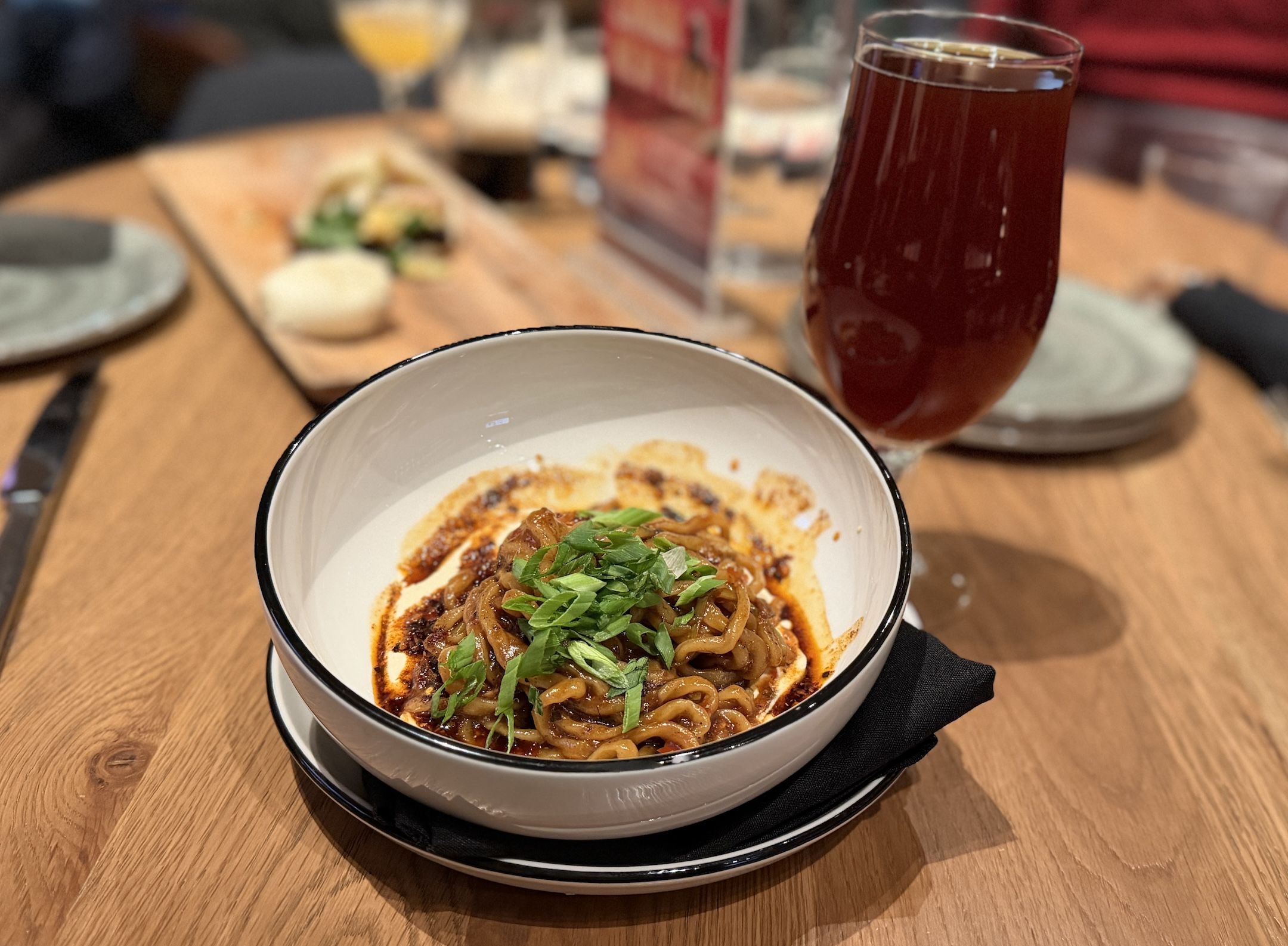 Bowl of noodles in spicy red sauce topped with chopped green onions, placed on a black napkin and white saucer on a wooden table next to a glass of dark amber beer.