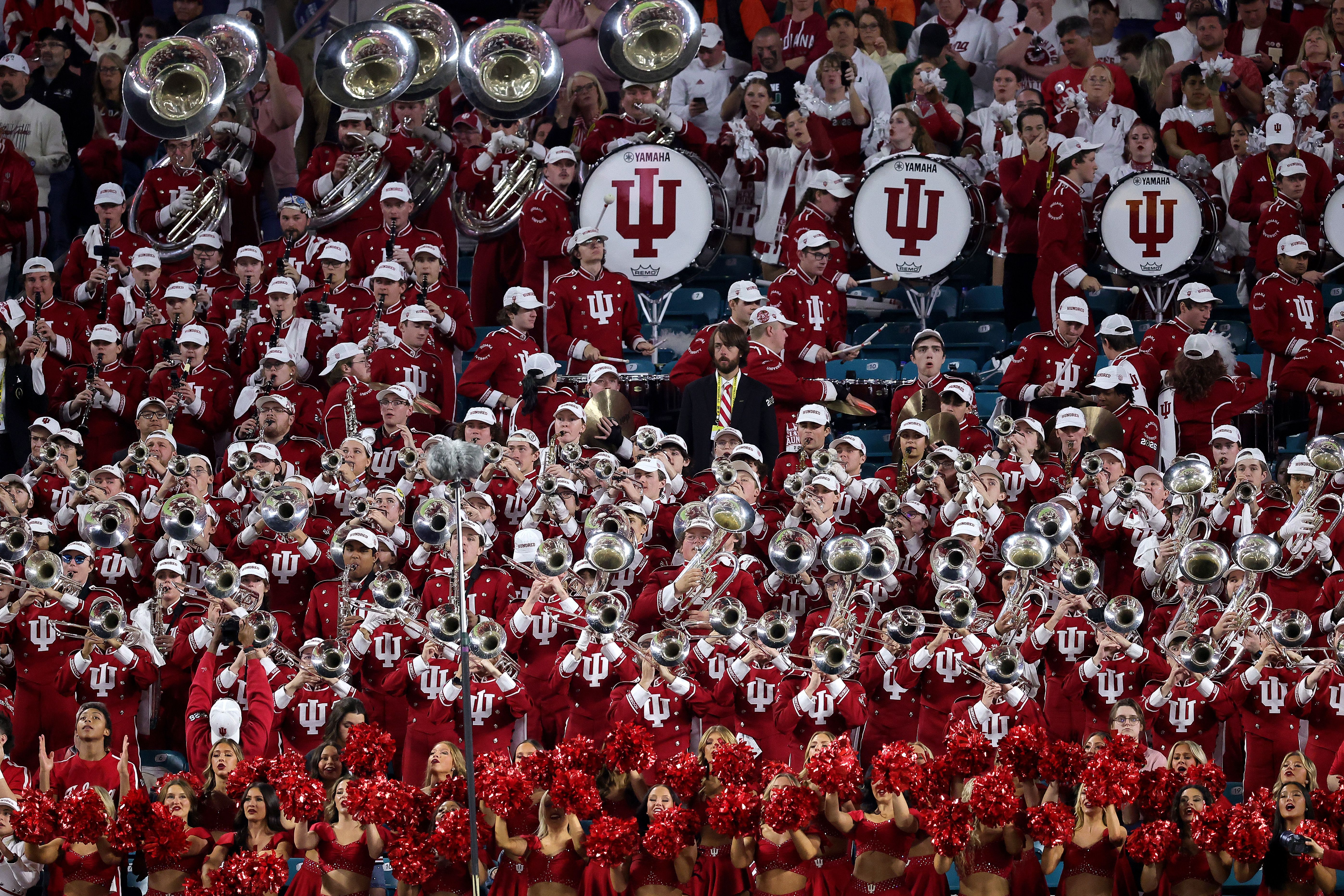 Indiana University marching band in red uniforms with white IU logos and white hats playing instruments, with cheerleaders in red outfits holding pom-poms in front, at a stadium event.