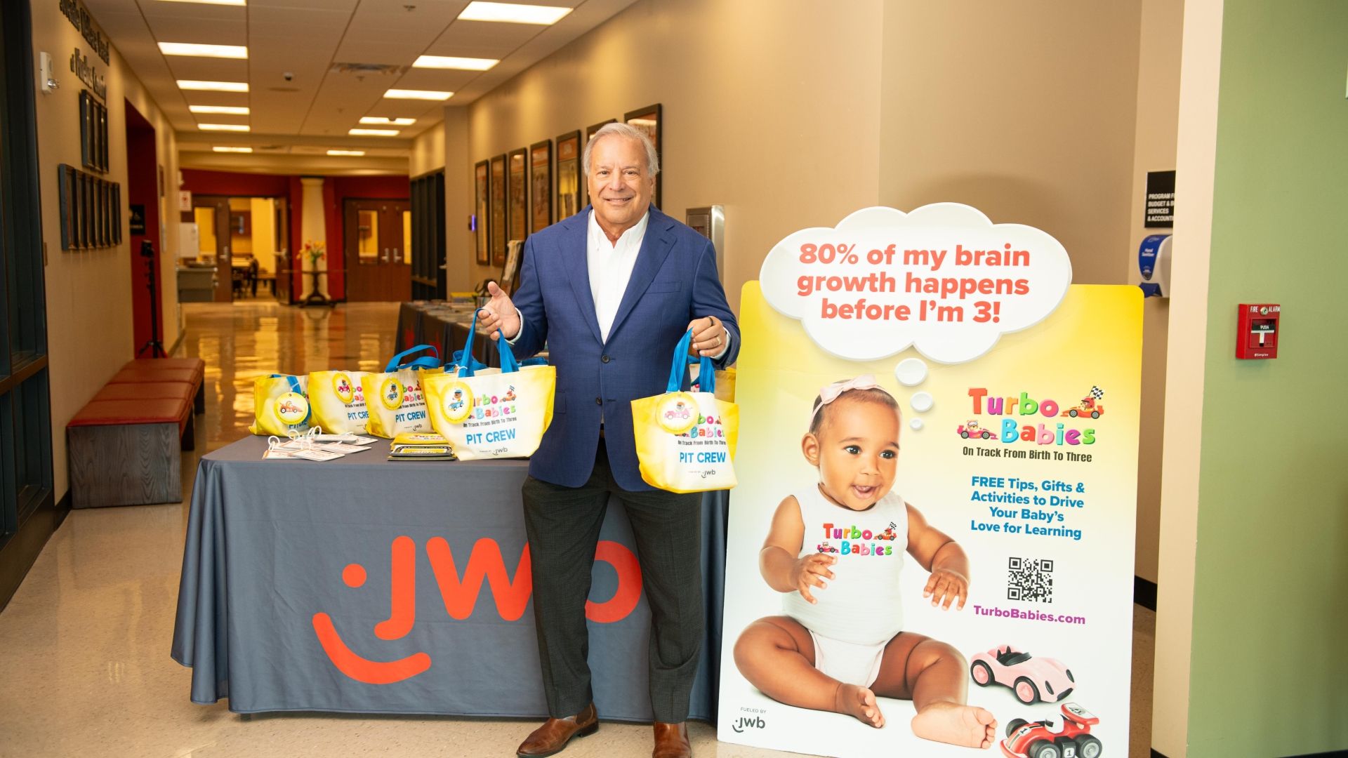 Man in blue blazer stands beside table with yellow Turbo Babies bags in hallway. Large Turbo Babies poster shows baby and text about brain growth before age 3.