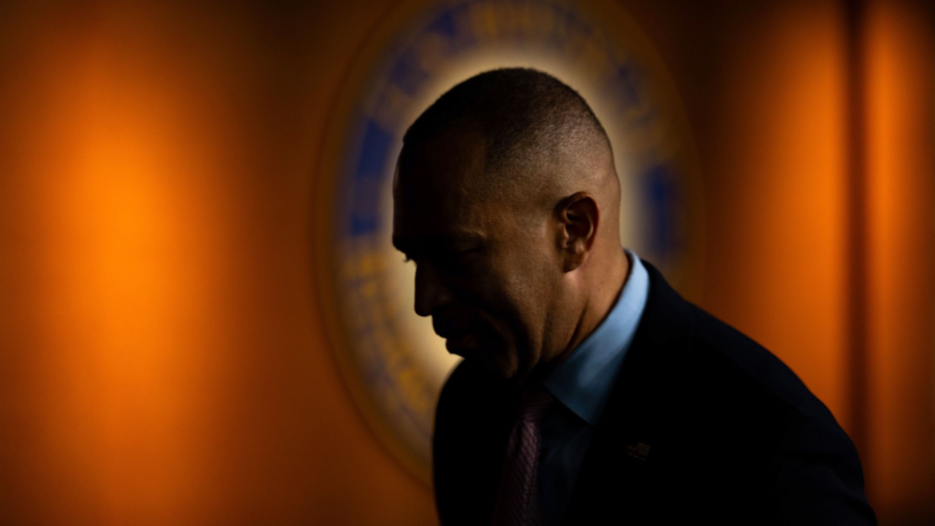 Minority Leader Hakeem Jeffries, wearing a dark suit and standing in front of a dimly lit House of Representatives seal.