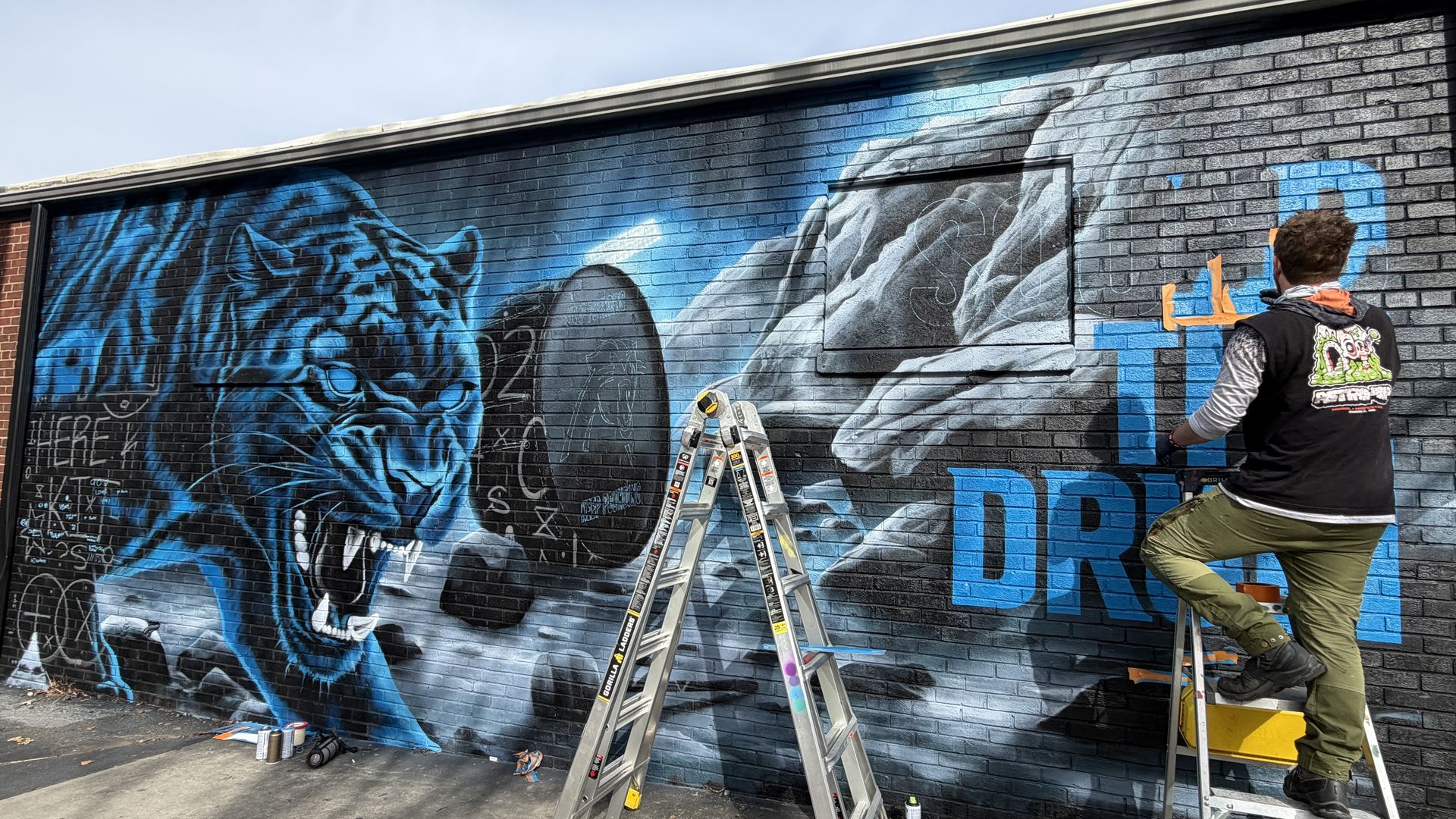 Artist painting a vibrant mural on a brick wall featuring a fierce blue tiger and large text starting with "THE DR" in blue, with ladders and paint cans on the ground nearby.
