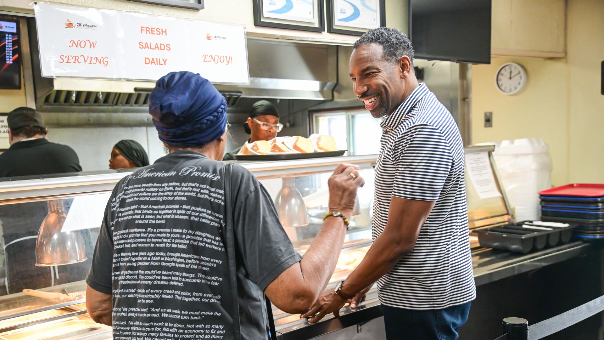 Two people interacting at a restaurant counter with food trays and signs above reading "Now Serving," "Fresh Salads Daily," and "Enjoy!"
