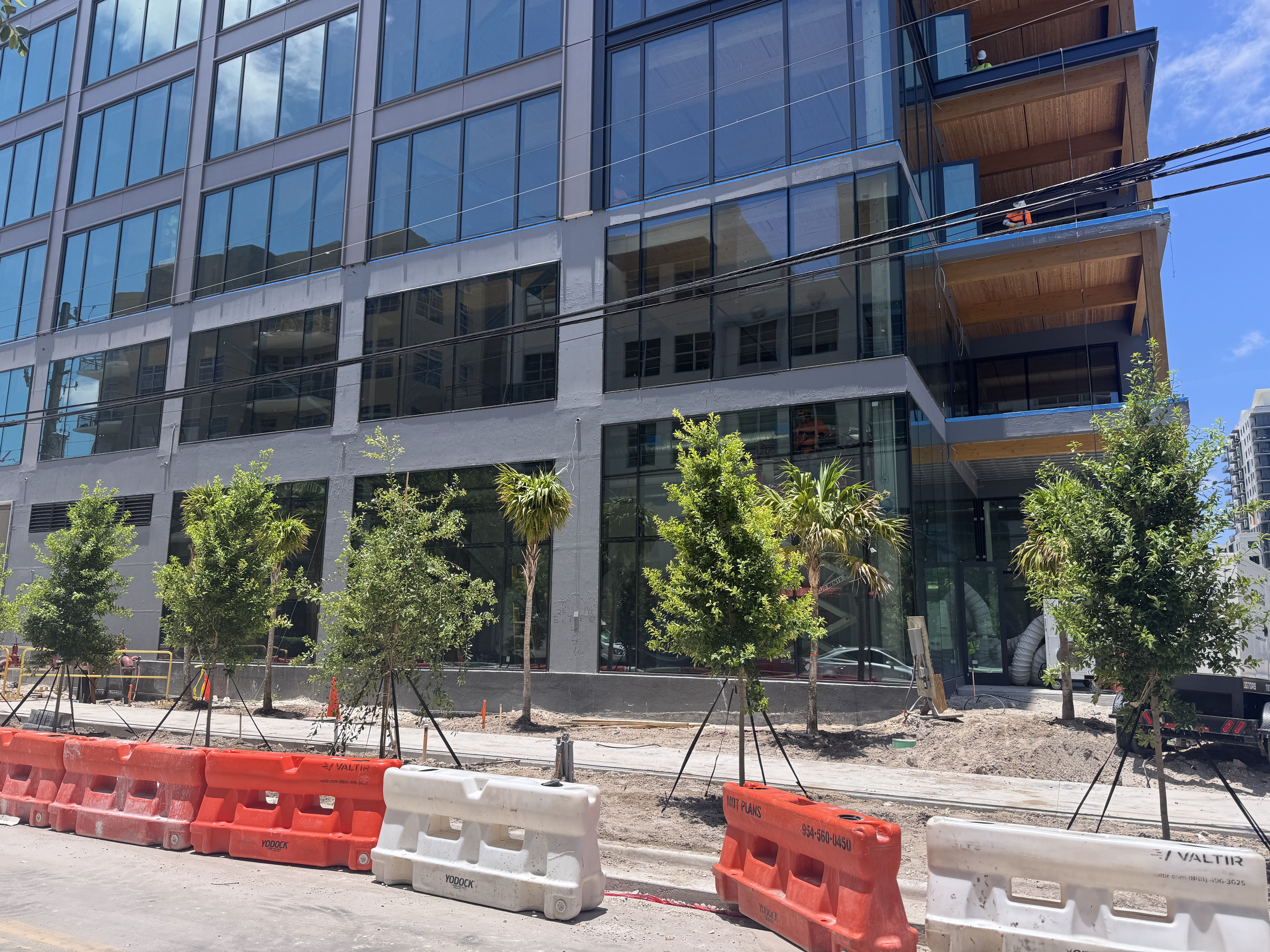 Modern glass-front office building under construction with wooden balconies on the right, workers in hard hats, orange and white barriers along the street, blue sky and trees in foreground.