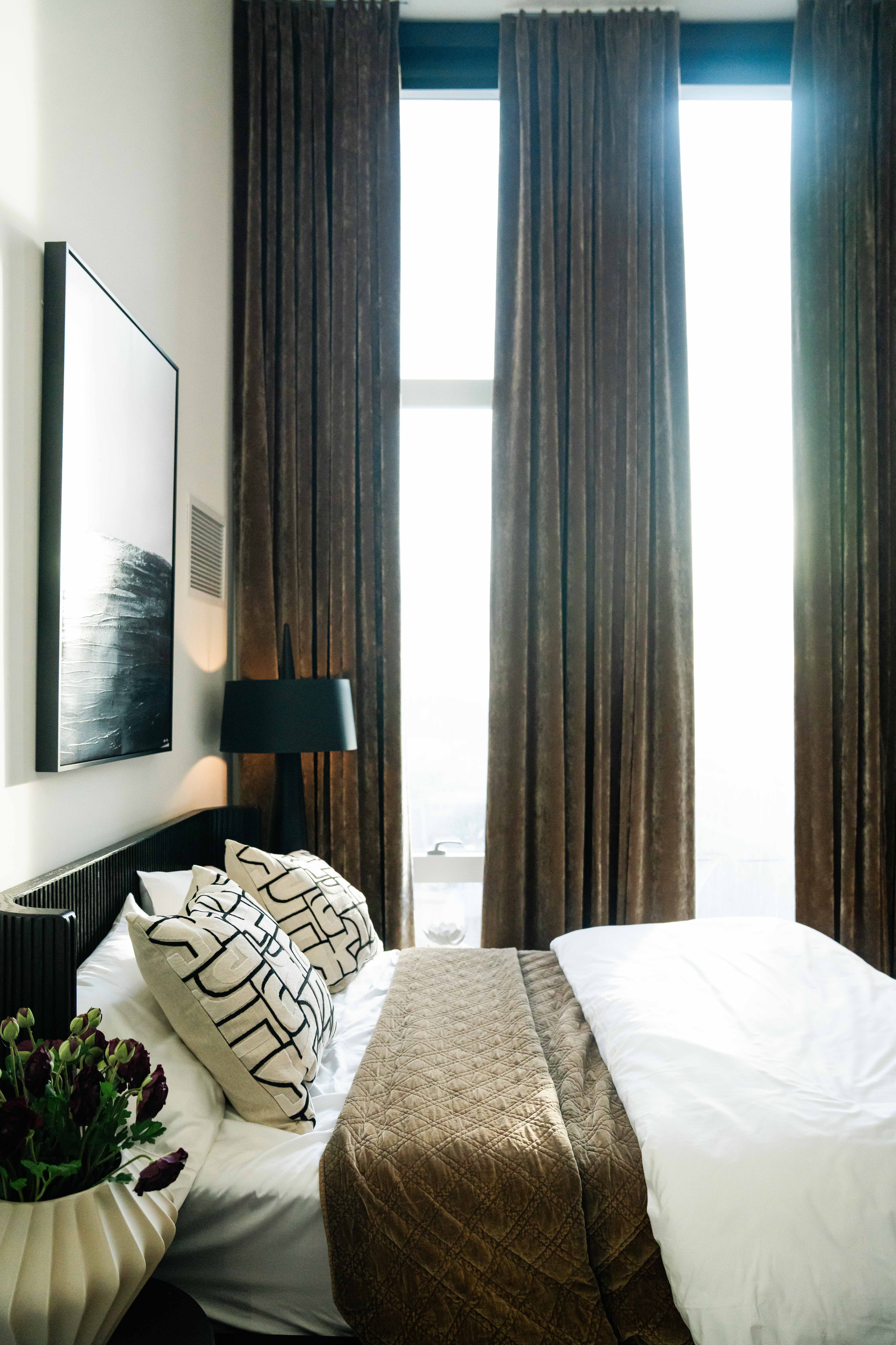 Bright bedroom with tall windows covered by brown curtains. Bed with white sheets, brown quilt, abstract patterned pillows. Black lamp and framed black-and-white artwork on wall. Vase with dark flowers.