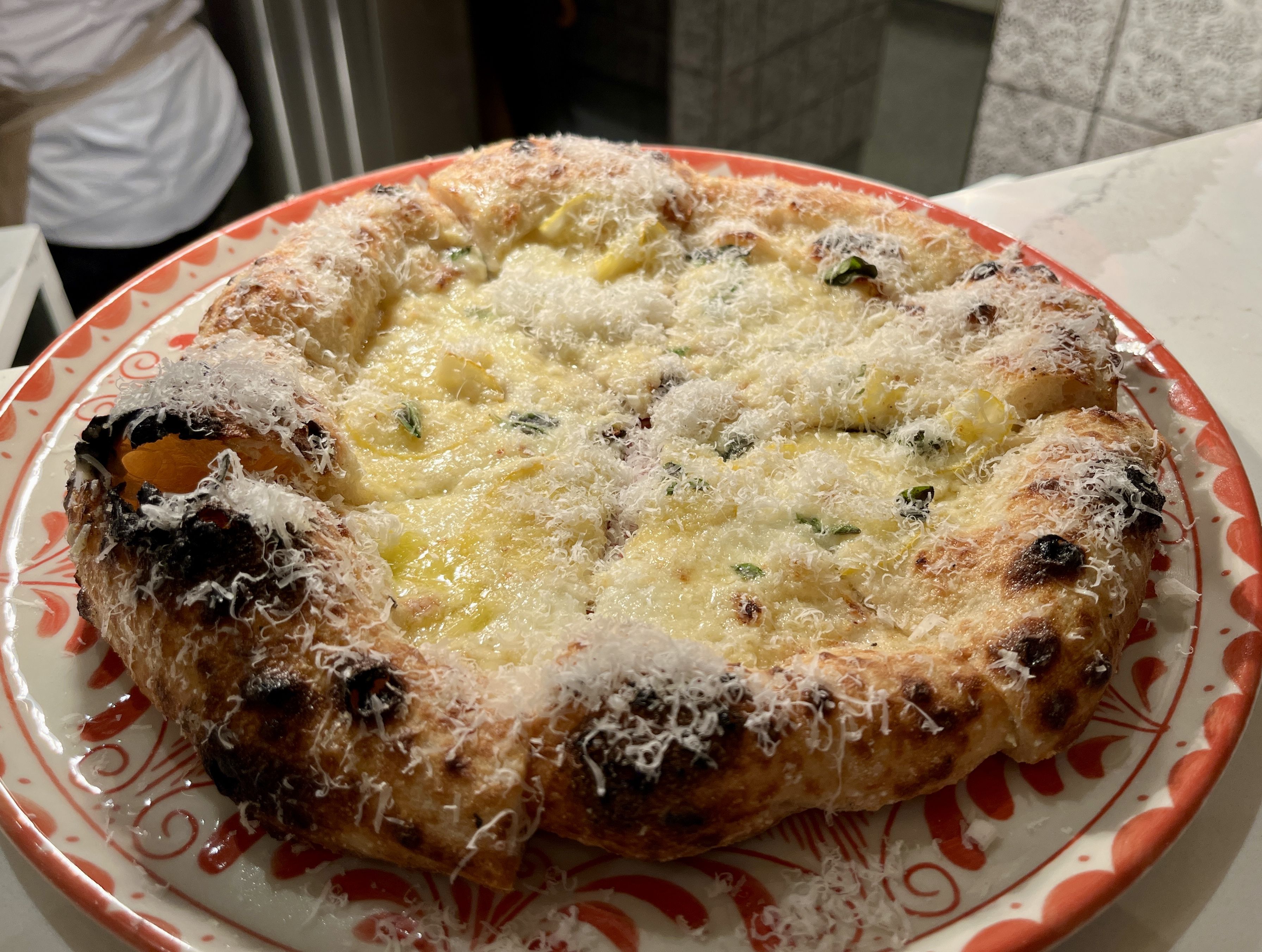 Close-up of a cheesy pizza with a thick, charred crust on a red and white patterned plate. The pizza is topped with melted cheese and sprinkled with grated cheese and herbs.