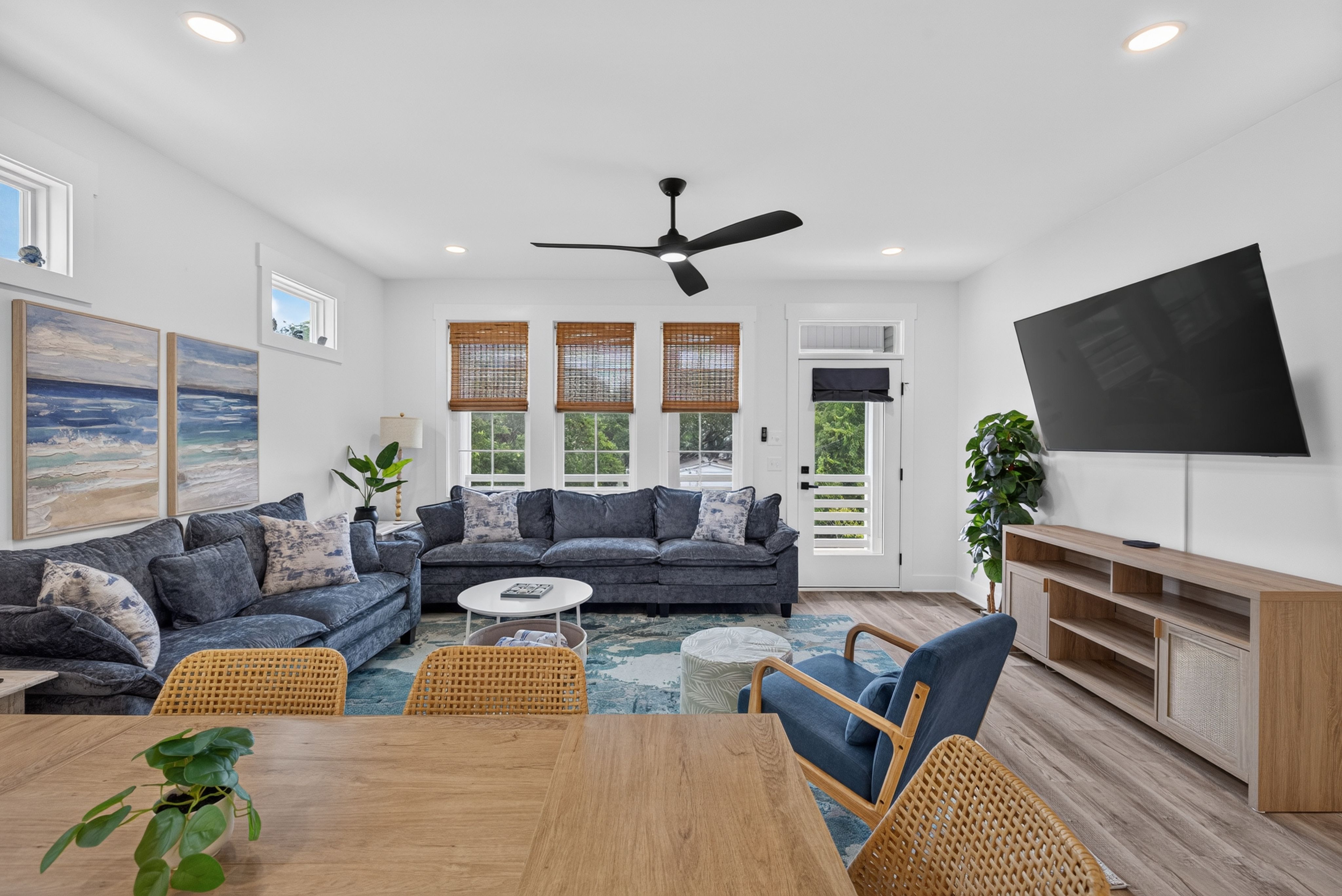 Bright modern living room with white walls, a dark blue sectional sofa, three windows with woven shades, a black ceiling fan, a large wall TV, and wooden tables with wicker chairs and green plants.