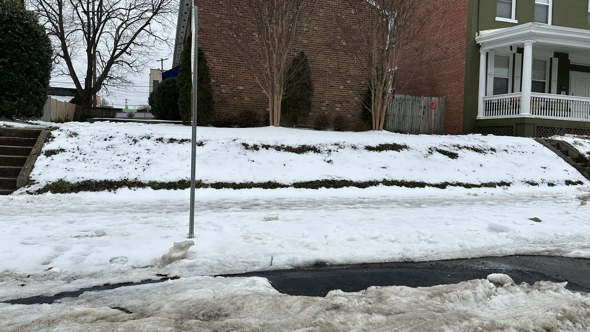 Snow-covered suburban street with a sloped front yard between a brick house and a green house with a white porch. Bare trees line the yard; a metal street sign pole stands in the foreground.
