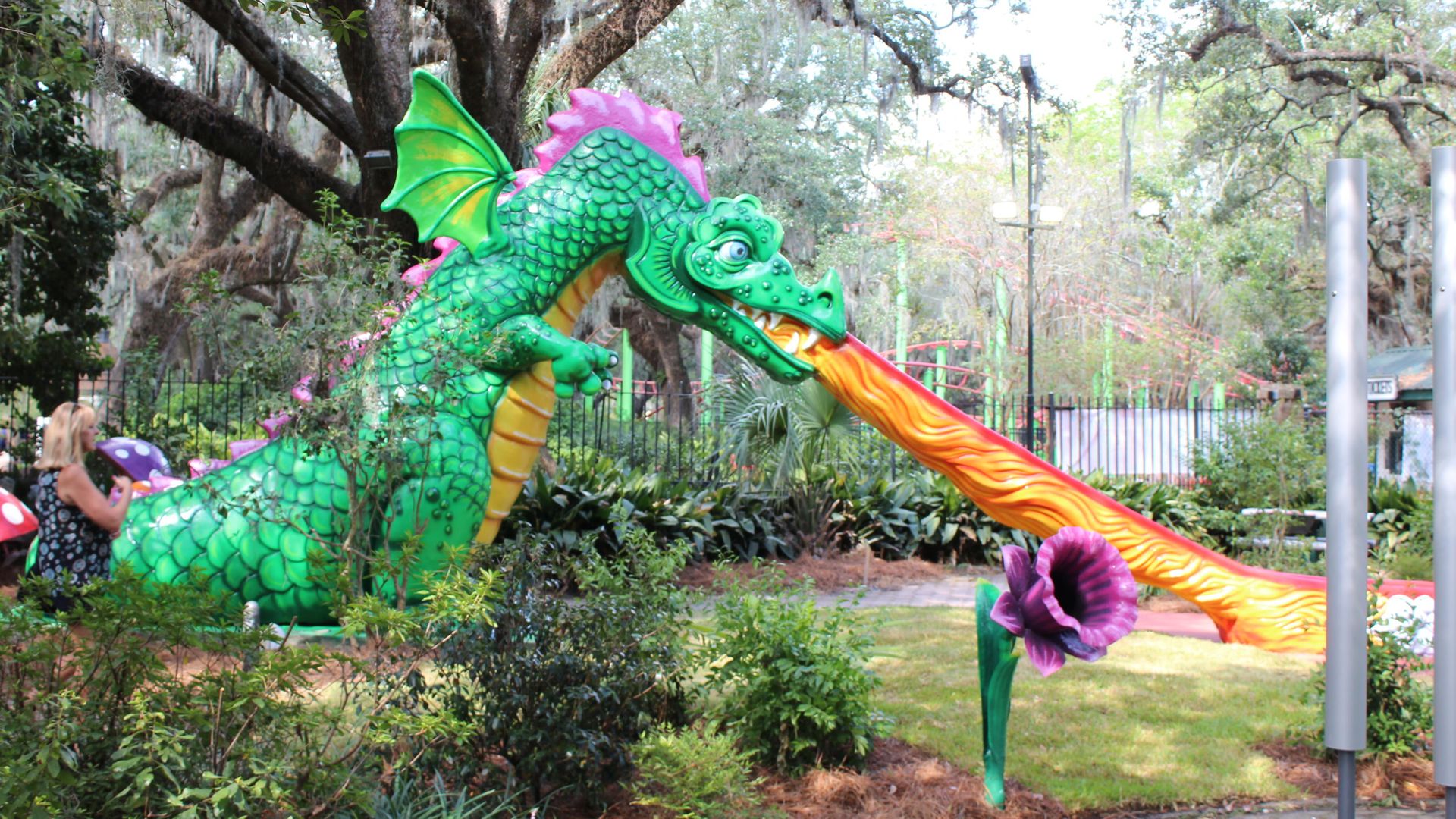 Green dragon statue with pink accents breathing a long stream of orange flames onto a purple flower in a garden with trees and a woman in a black dress nearby.