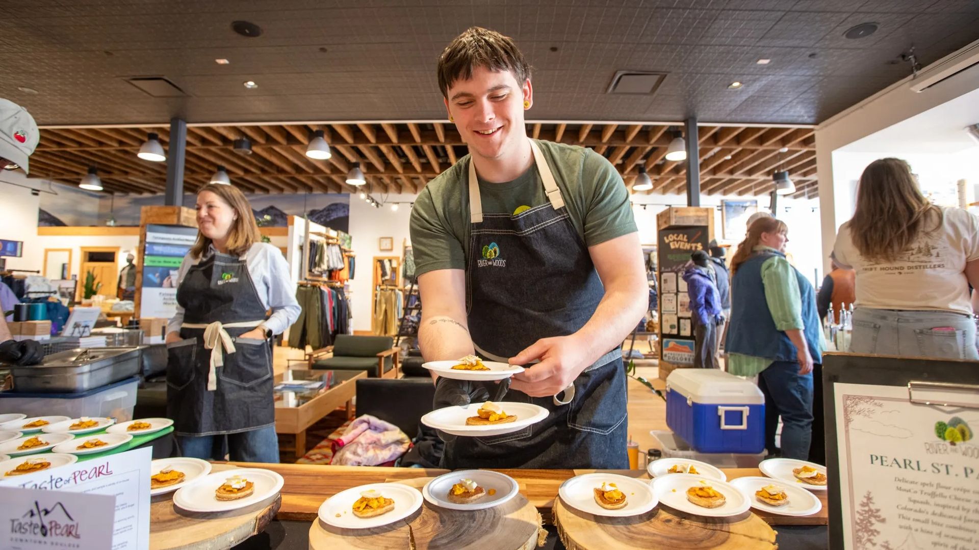 Young man in a green t-shirt and dark apron stacks small plates of food on wooden rounds at a busy indoor tasting, with staff and shoppers visible in the background.