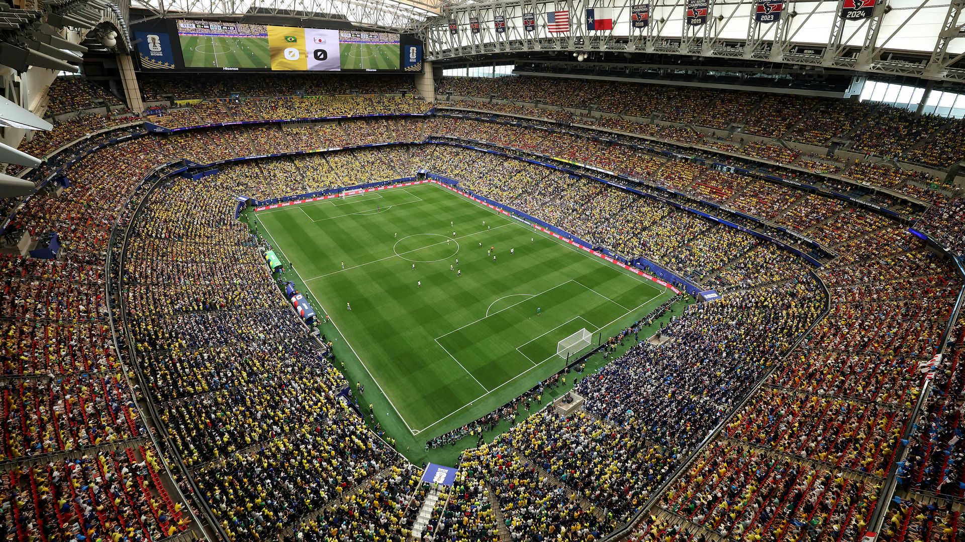 Wide panoramic view inside a crowded stadium with a green soccer pitch, a large video screen displaying a match, and fans in yellow and red seats. Visible flags and banners hang from the upper rim.