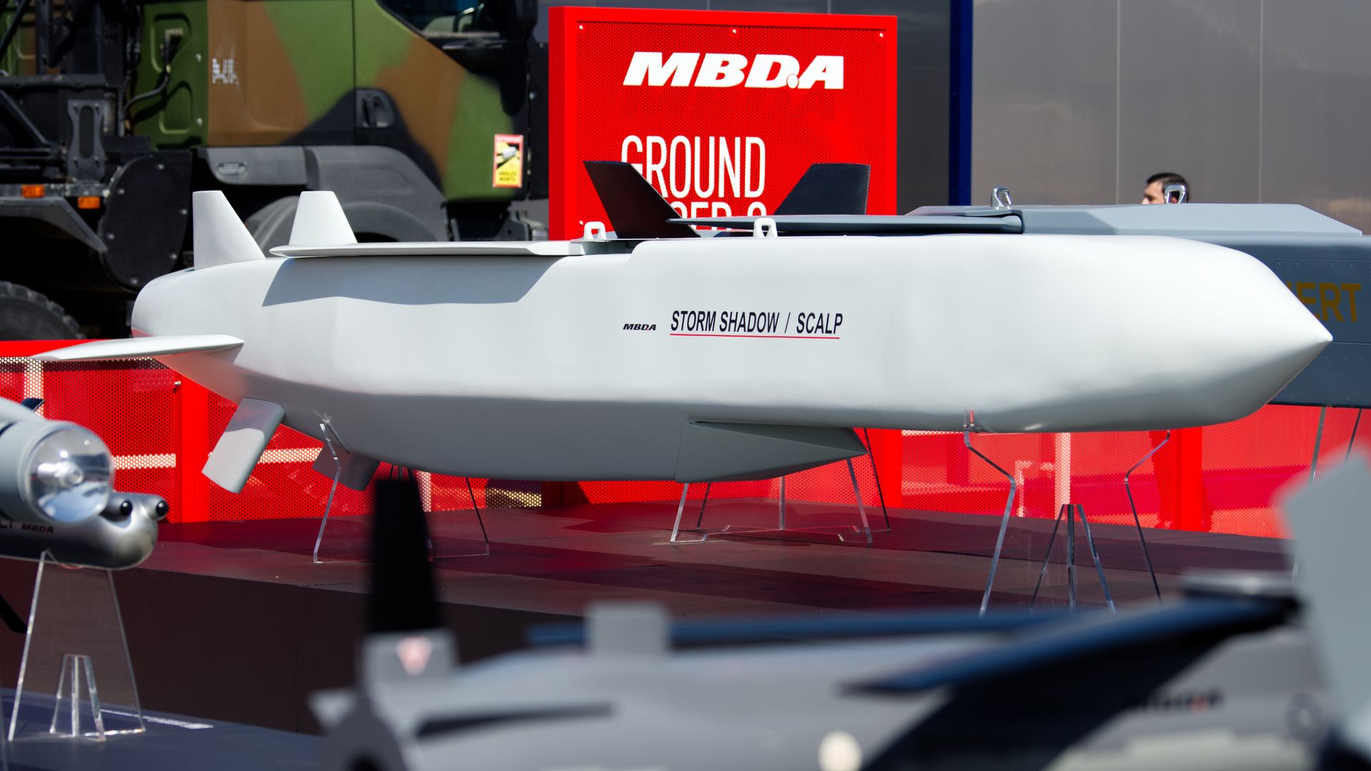 Models of missiles are on display at an outdoors defense-conference booth. A large red sign in the background reads: "MBDA."